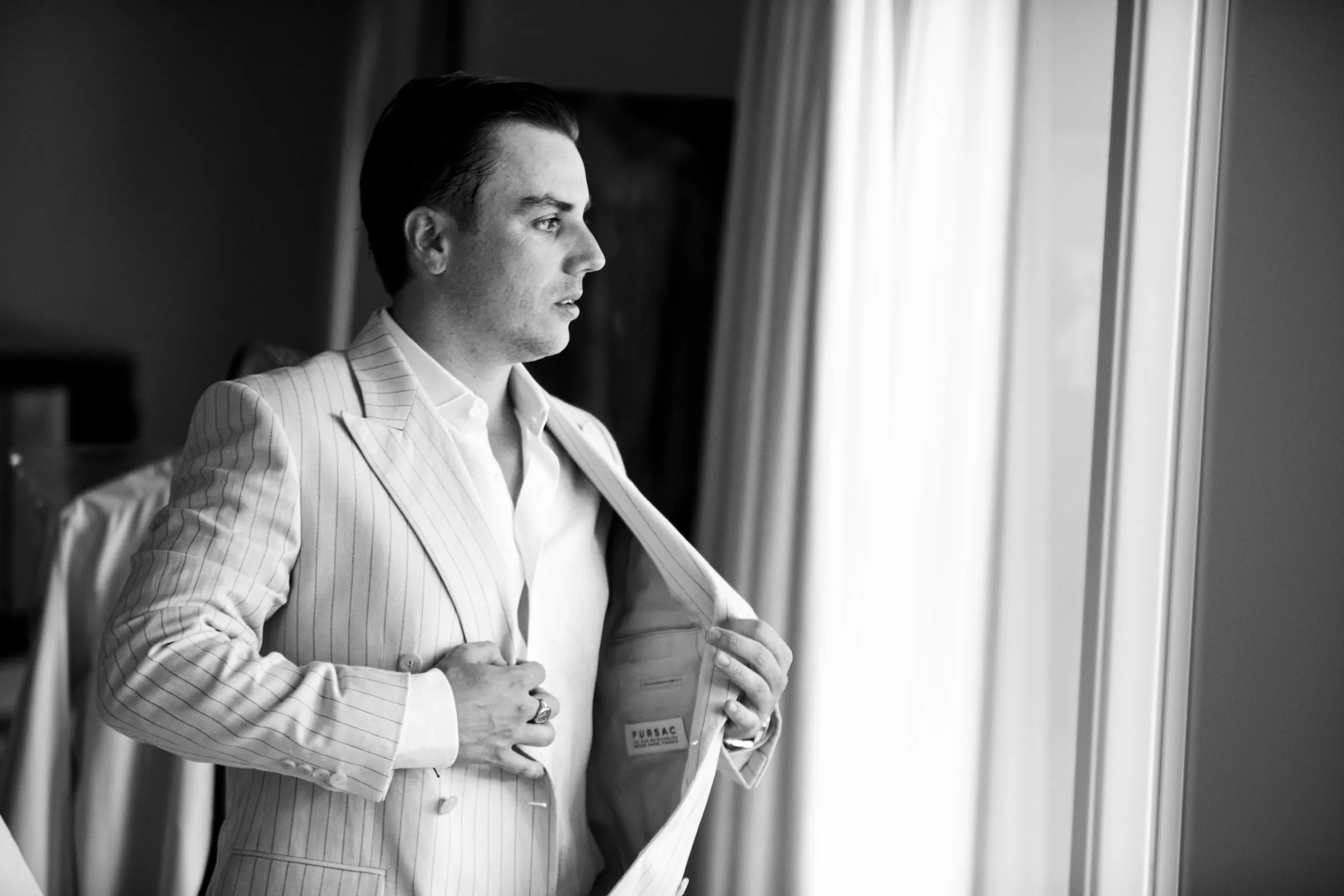 Black and white of the groom putting on his striped jacket by the window, Barbillón, Marbella