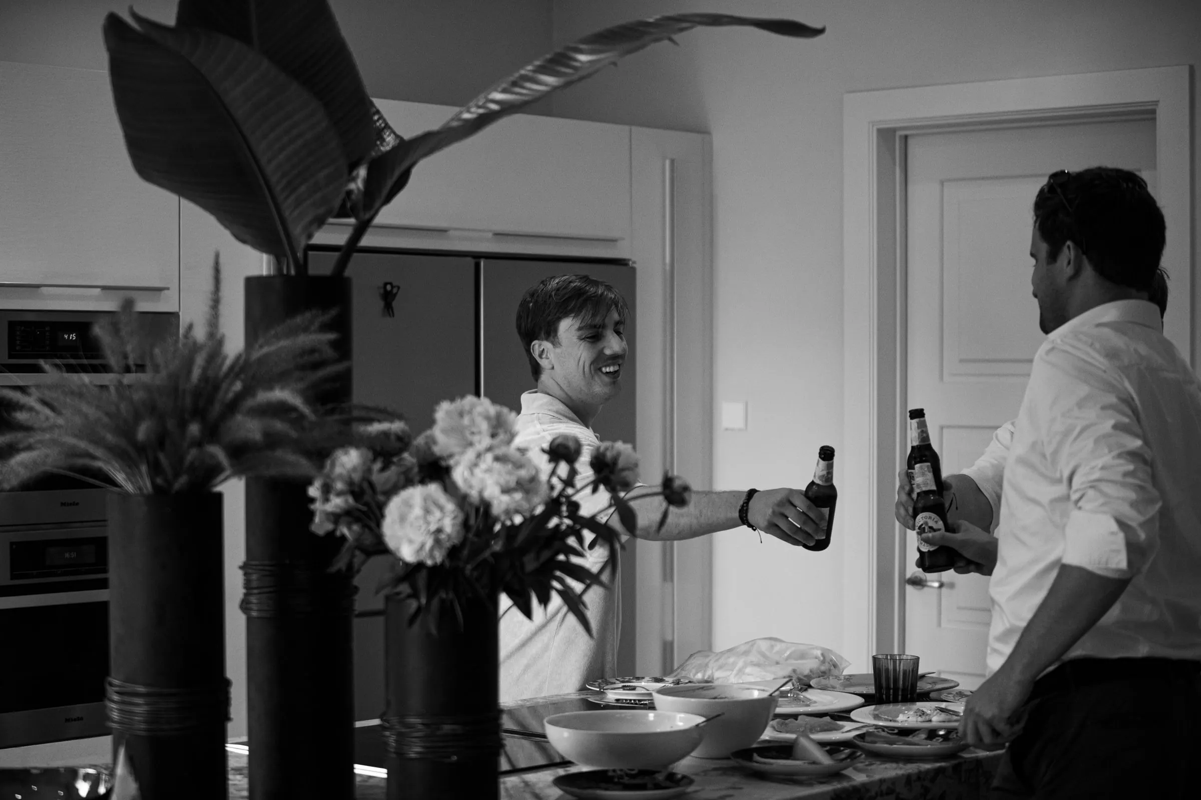 Black and white of the groom and a friend clinking beer bottles in the kitchen, Barbillón, Marbella