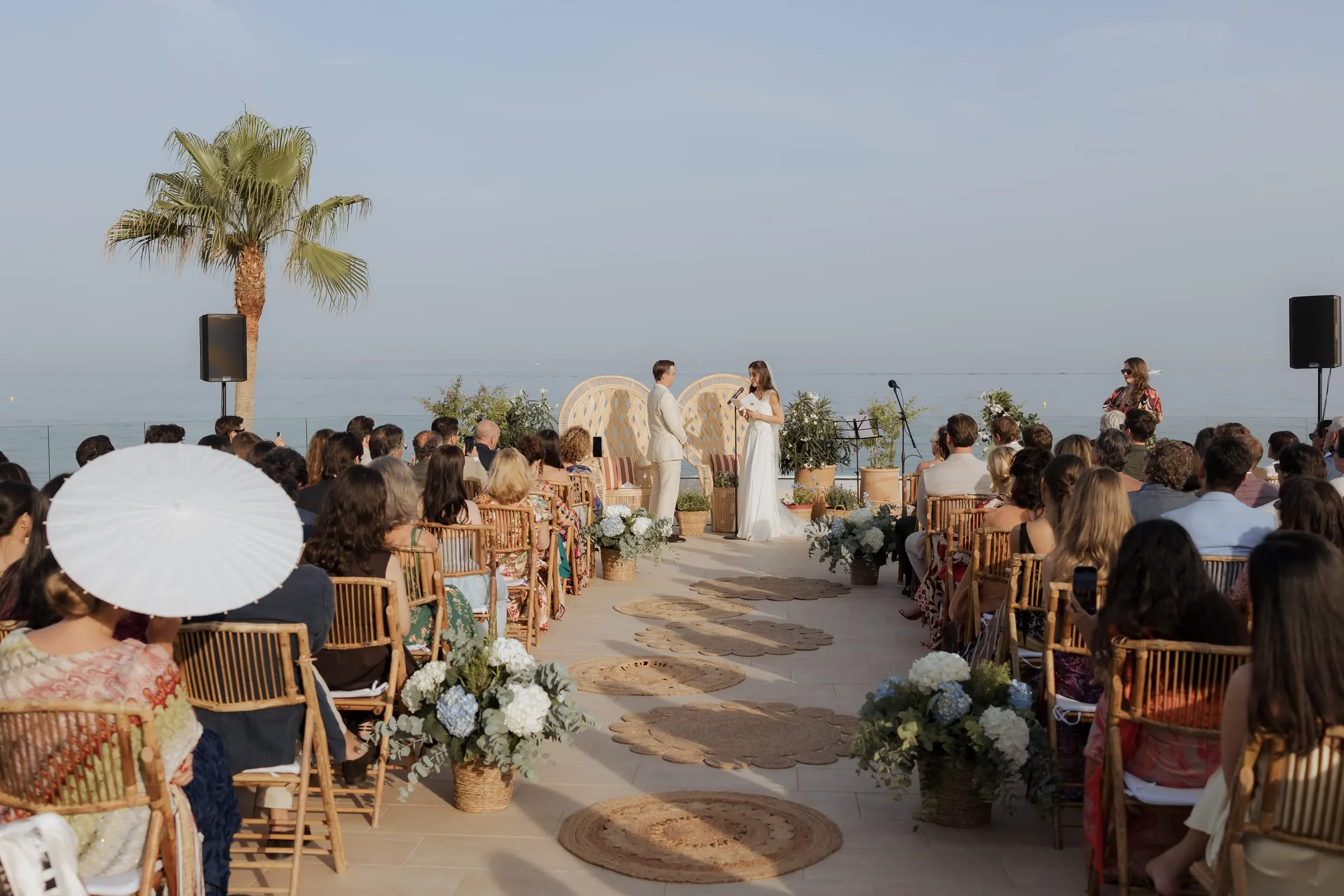 Seafront ceremony on the terrace seen from behind the guests, couple between wicker chairs, Barbillón, Marbella