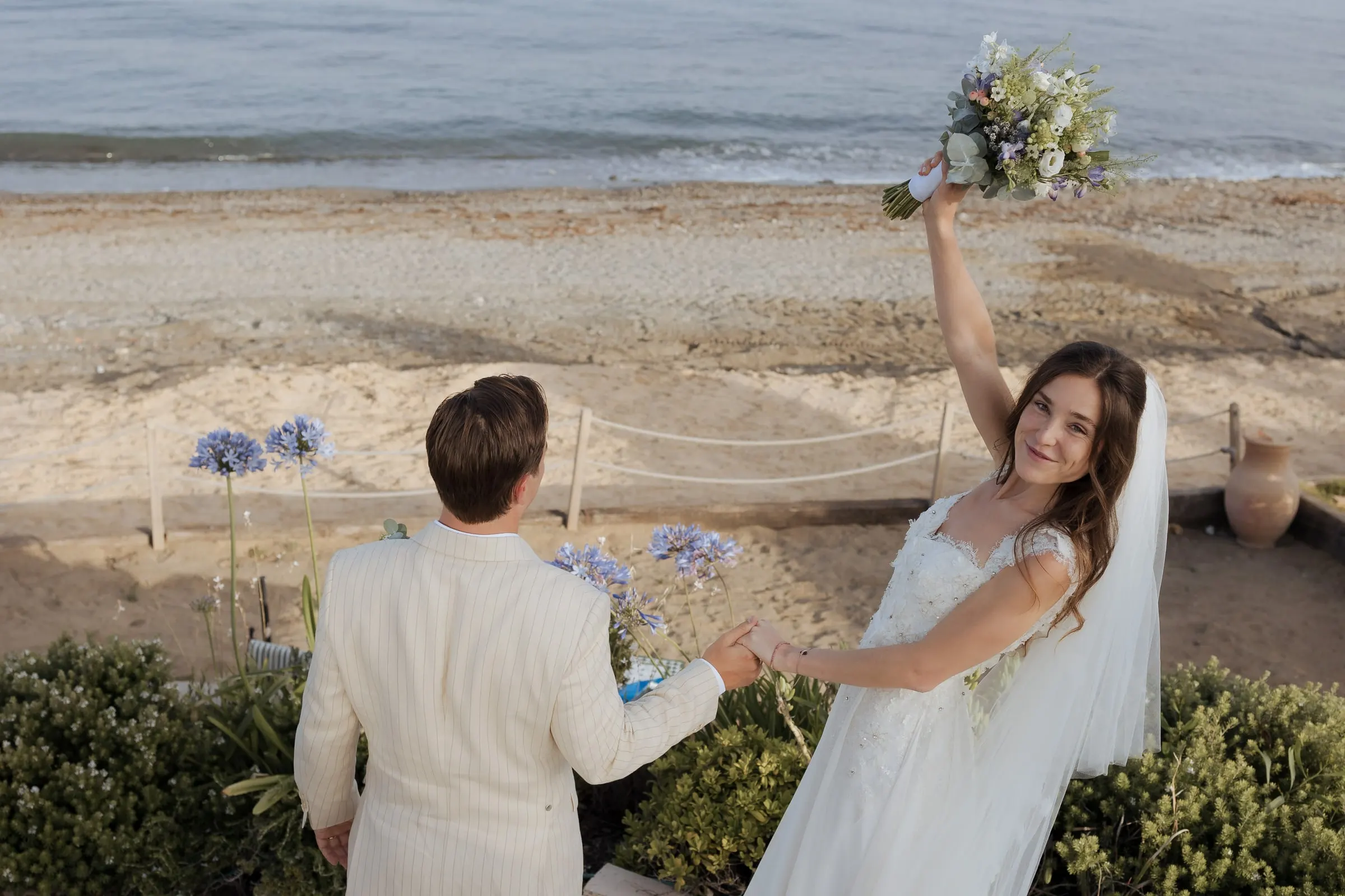 Bride raising her bouquet and holding the groom's hand on a path by the beach at Barbillón, Marbella