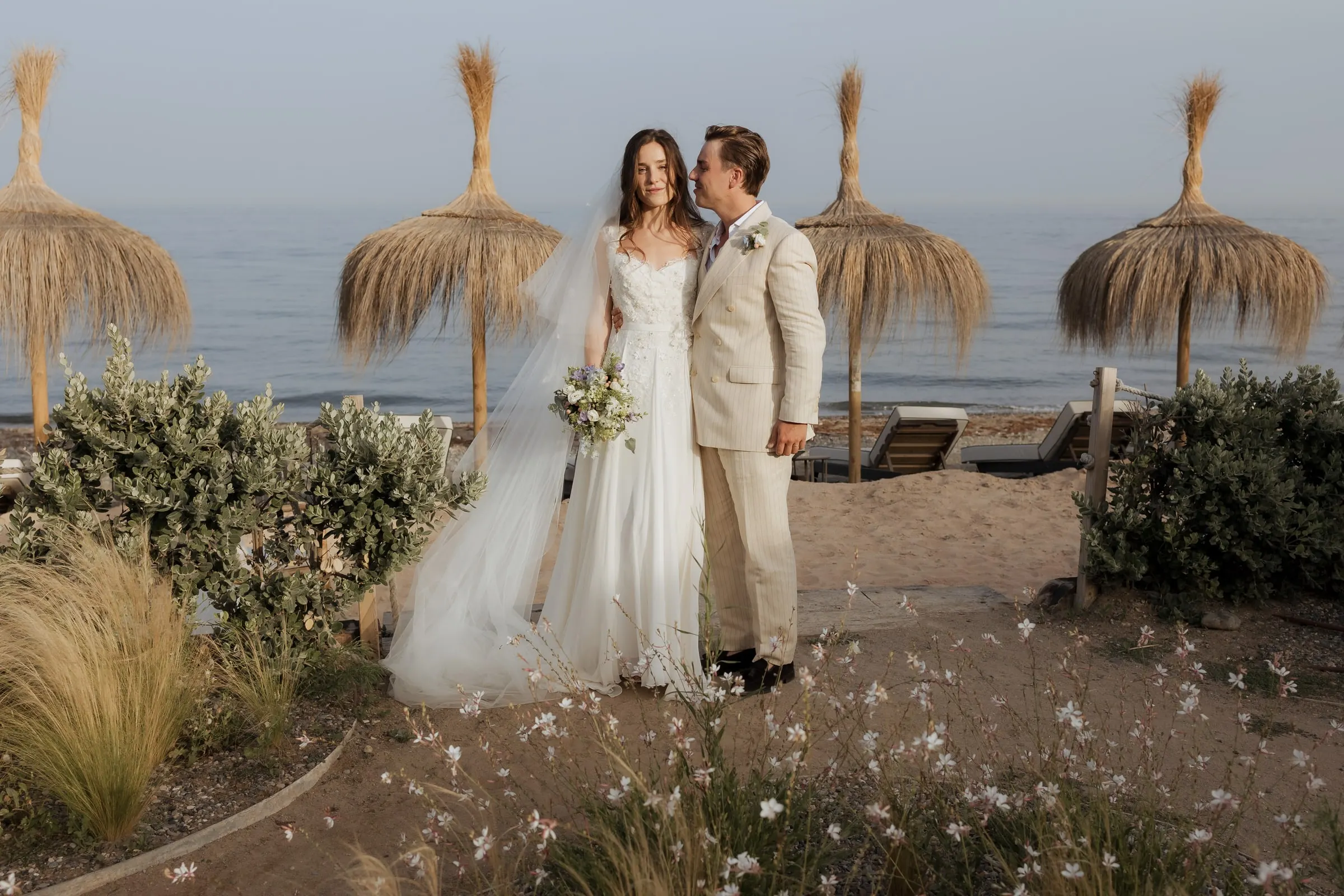 Bride and groom standing among dune plants and straw beach umbrellas by the sea at Barbillón, Marbella