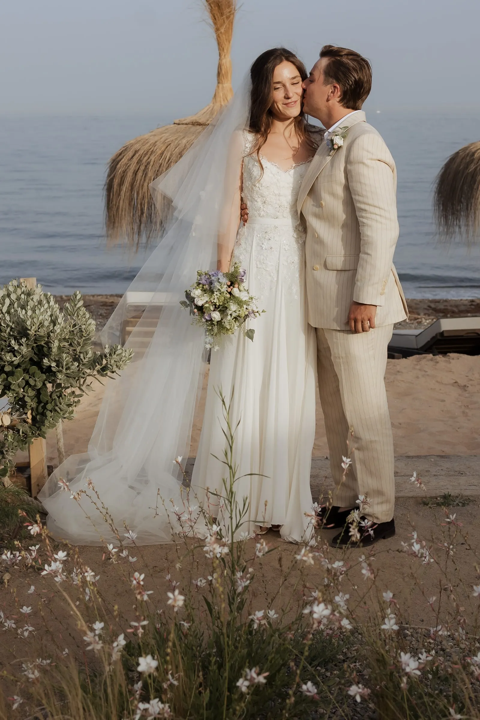 Groom kissing the bride's cheek among wild flowers and a straw beach umbrella at Barbillón, Marbella