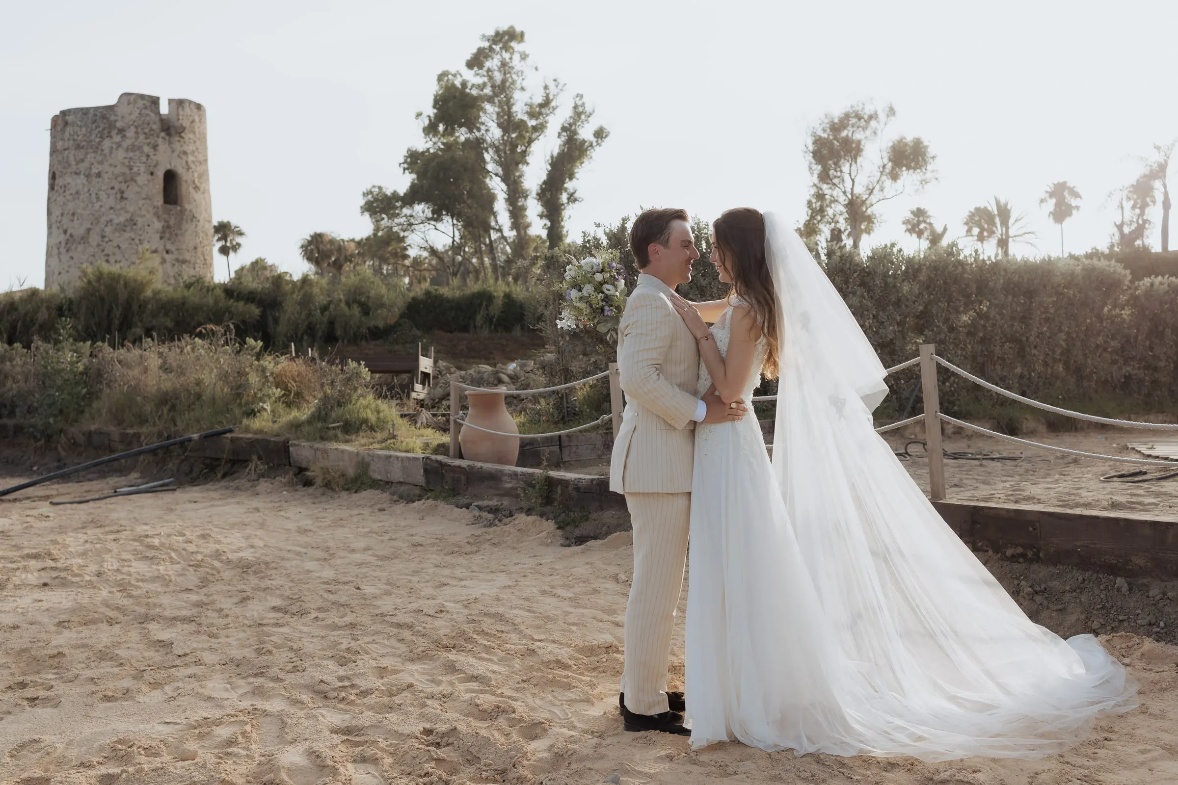 Bride and groom facing each other on the sand with an old stone watchtower behind, near Barbillón, Marbella