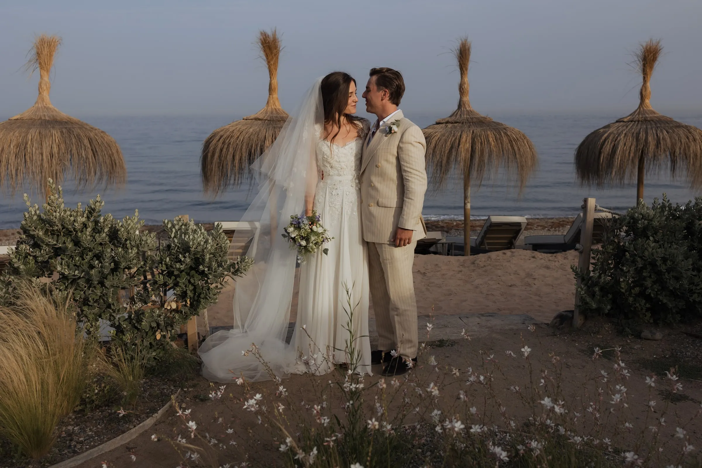 Bride and groom among dune plants and straw parasols by the beach, Barbillón, Marbella