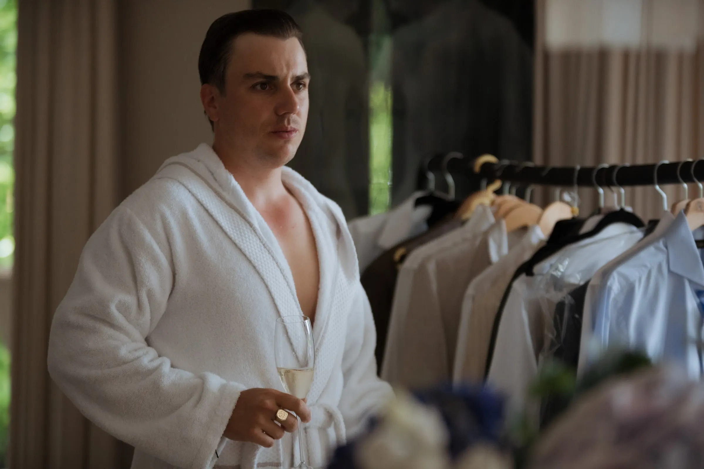 Groom in a white bathrobe holding a glass by a rail of suits, Barbillón, Marbella