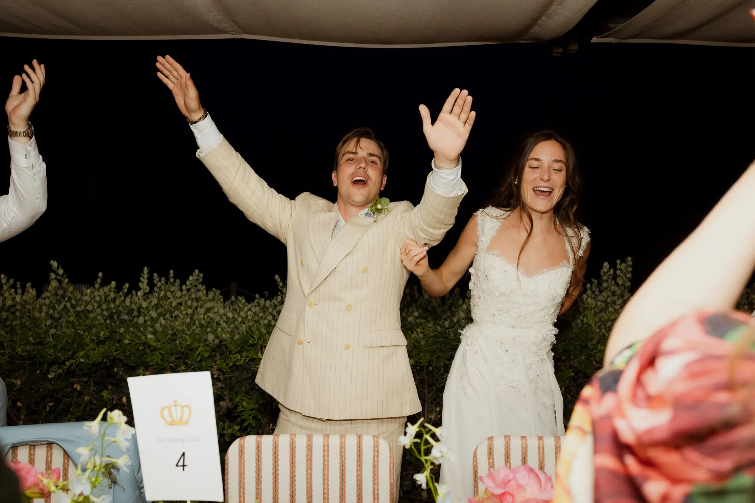 Bride and groom cheering with arms raised at the dinner table at night, Barbillón, Marbella