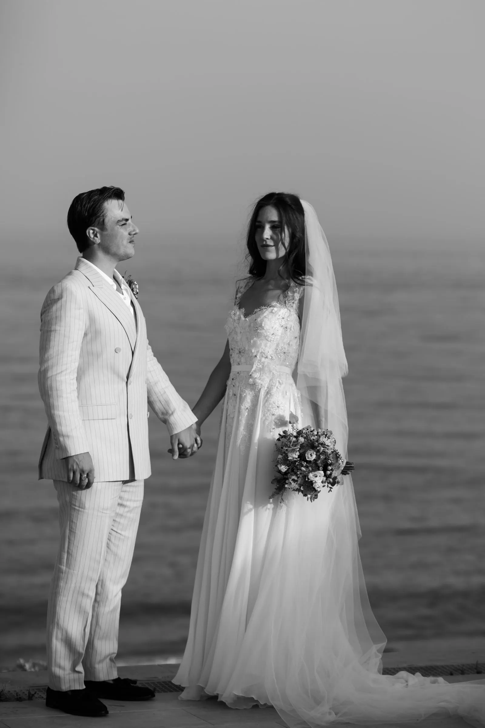 Black and white of the couple holding hands and smiling with the sea behind, Barbillón, Marbella