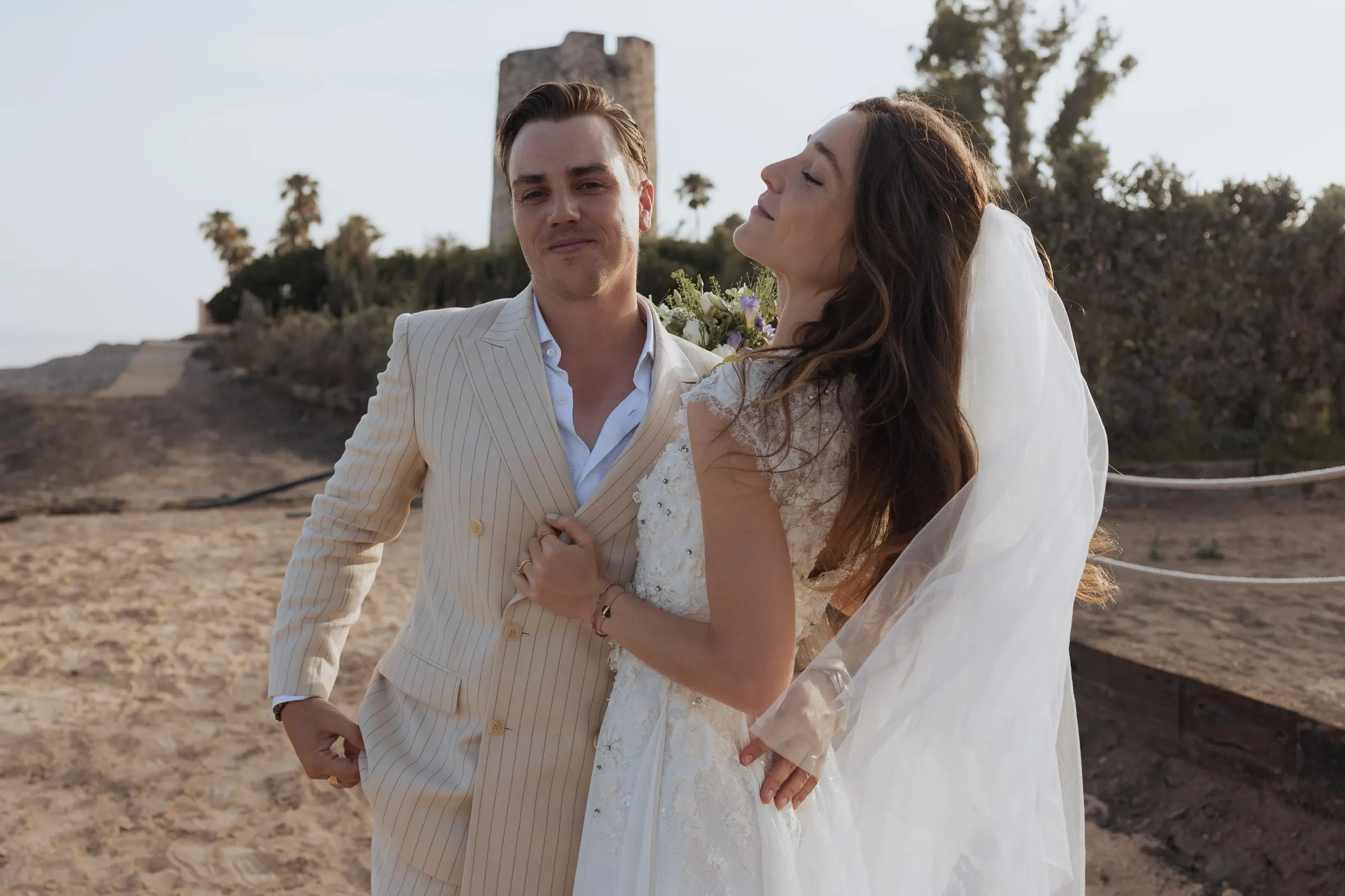 Bride tilting her head back laughing beside the groom near the watchtower, Barbillón, Marbella