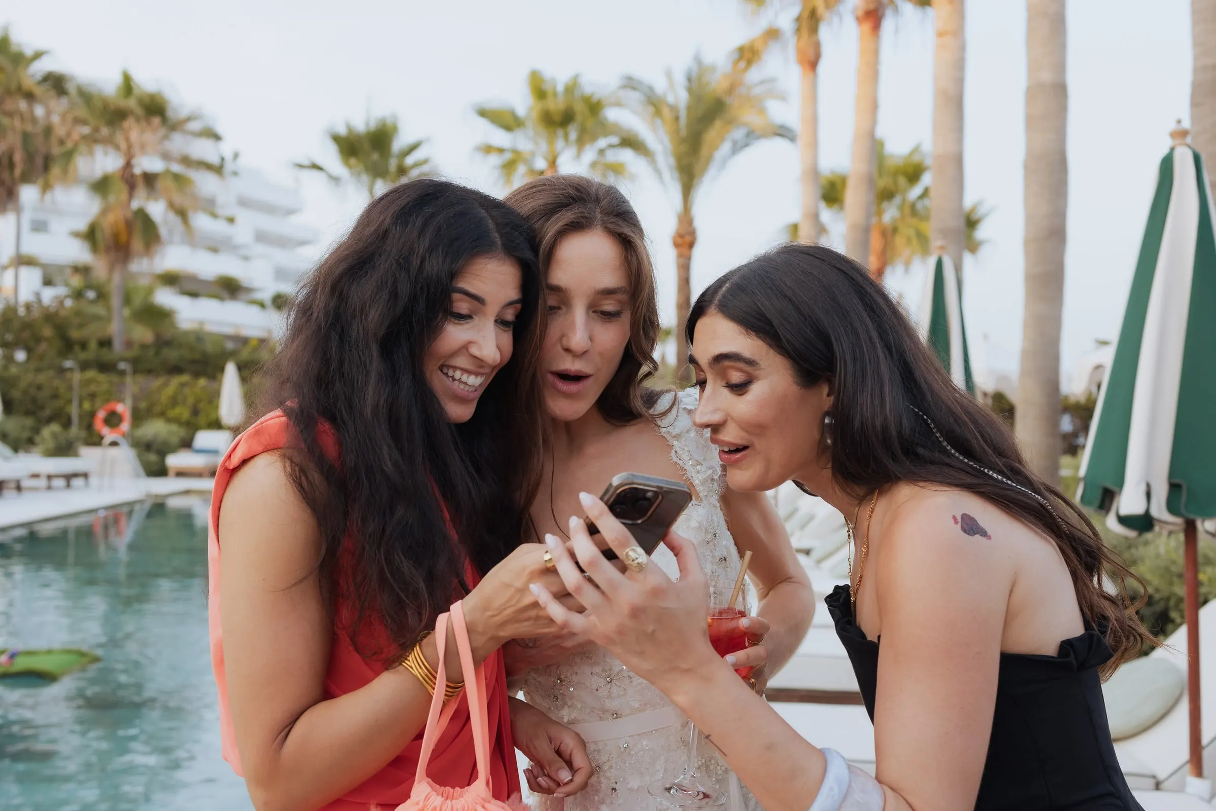 Bride and two friends laughing at a phone screen by the pool, Barbillón, Marbella