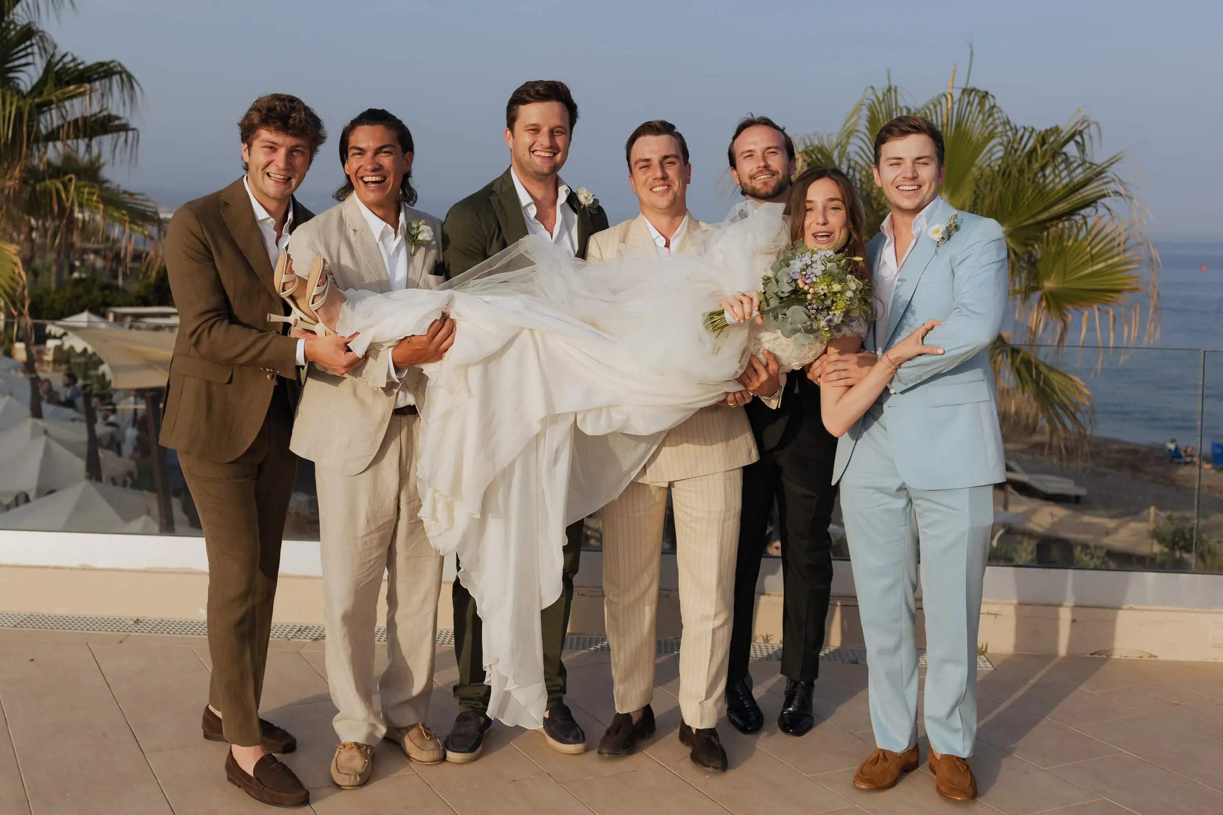 Groomsmen in colourful suits lifting the bride on the terrace above the sea, Barbillón, Marbella