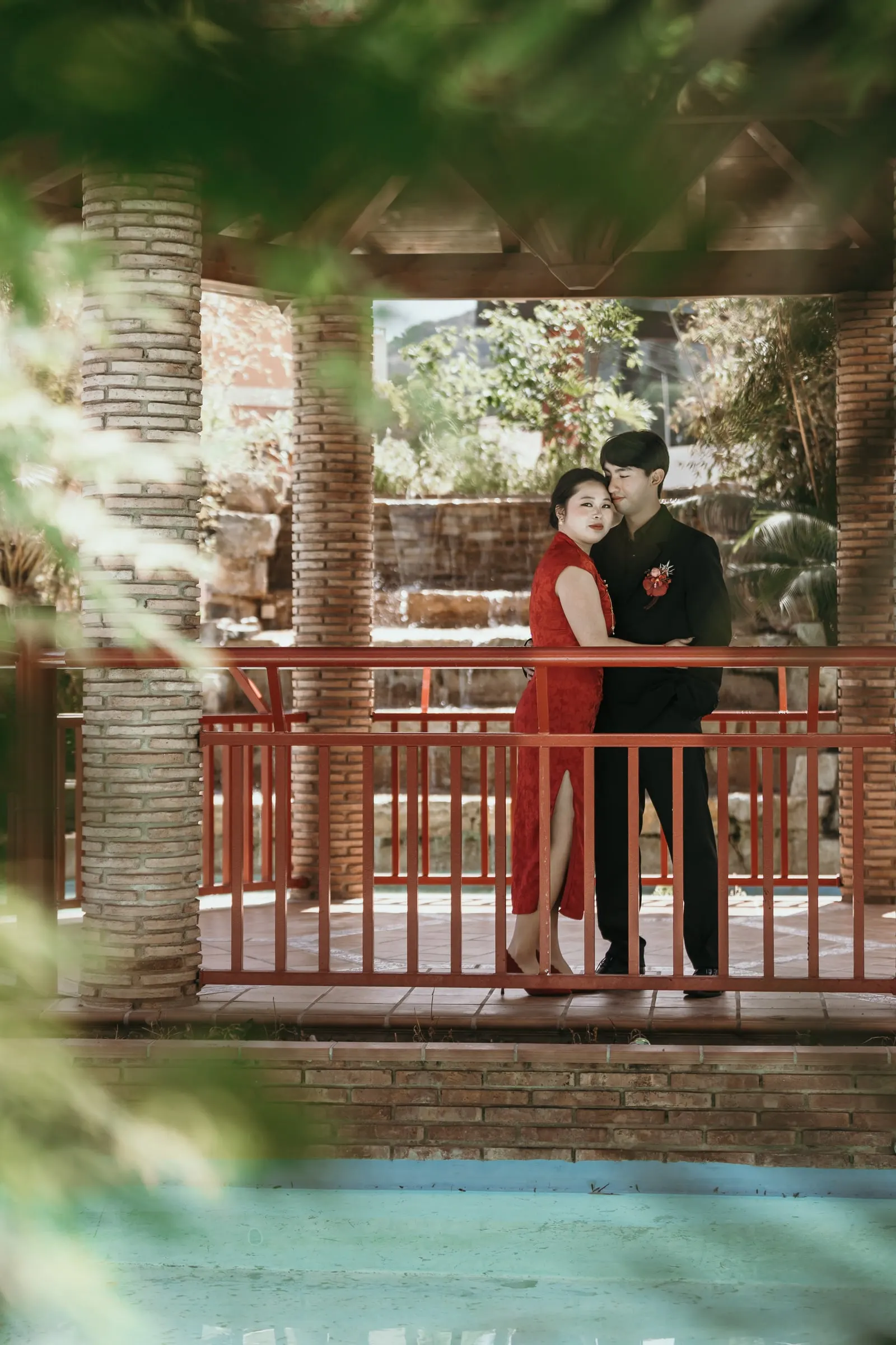 Couple embracing on a red bridge, seen between the pavilion pillars and the leaves, Jardín Oriental Bienquerido