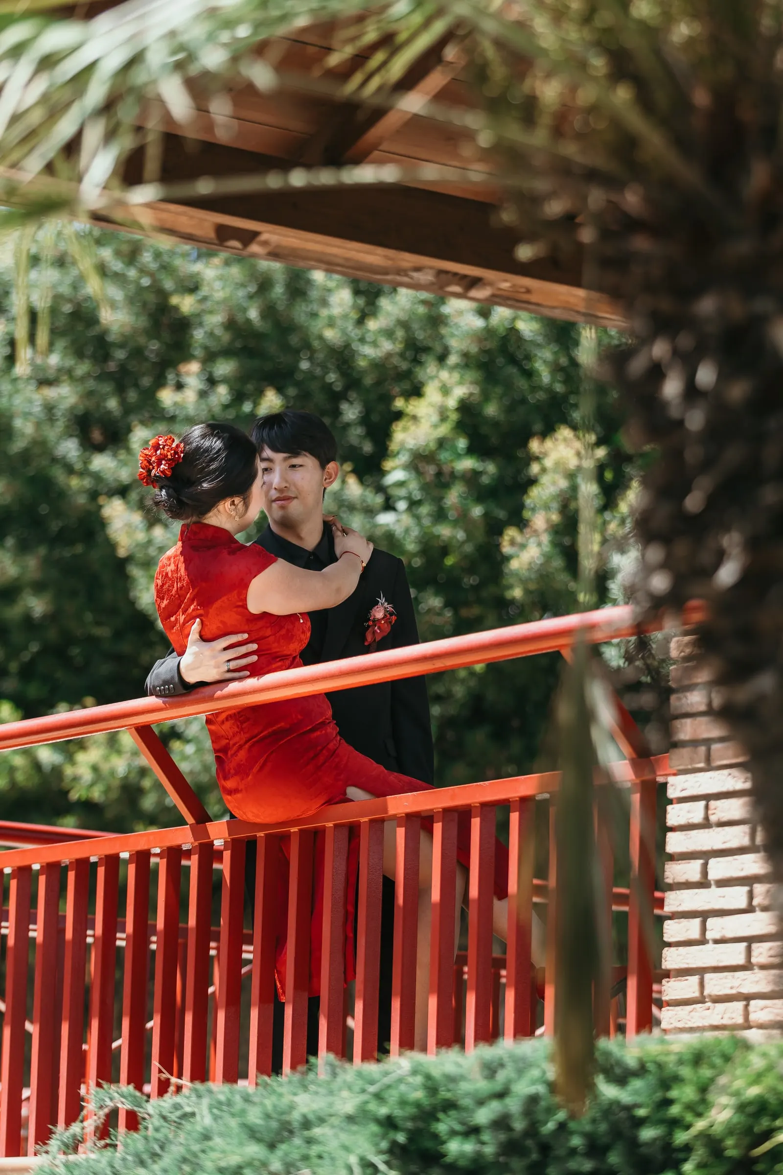 Bride-to-be in her red qipao embracing her fiancé at the pagoda railing, framed by palm leaves
