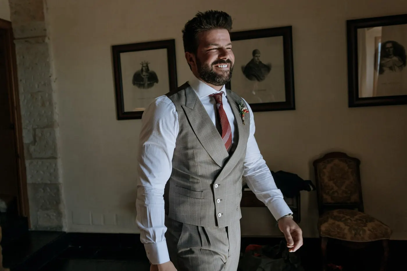 Groom ready in his suit, smiling in a room hung with old prints, Castillo del Buen Amor