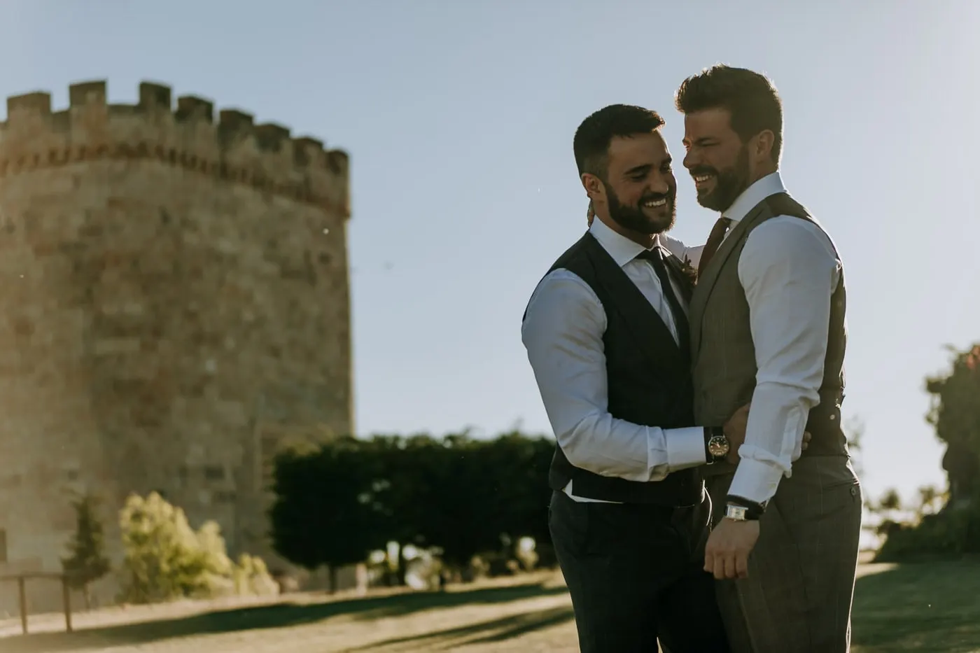 The two grooms laughing in an embrace beside the round tower of Castillo del Buen Amor, Salamanca