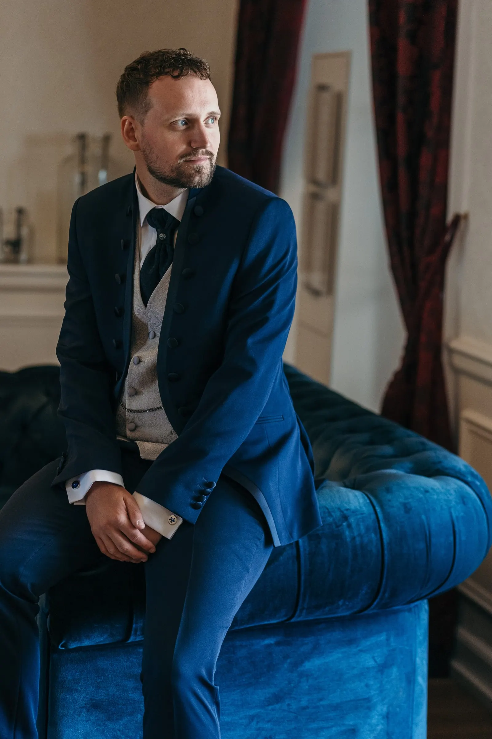 Vincent in a navy suit sits on a blue velvet sofa in his hotel room before the wedding
