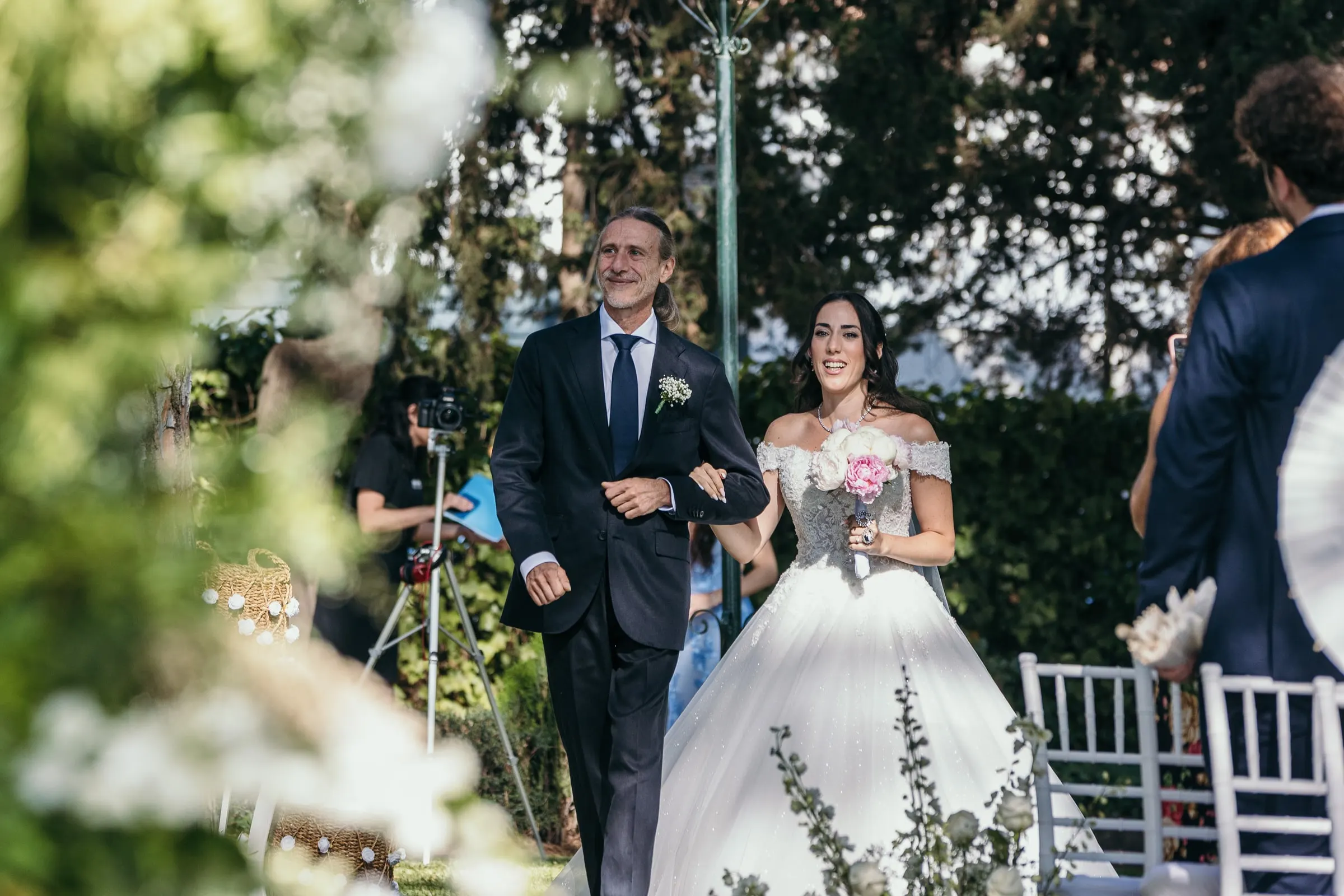 Sara is walked down the aisle between white flowers by a man in a navy suit