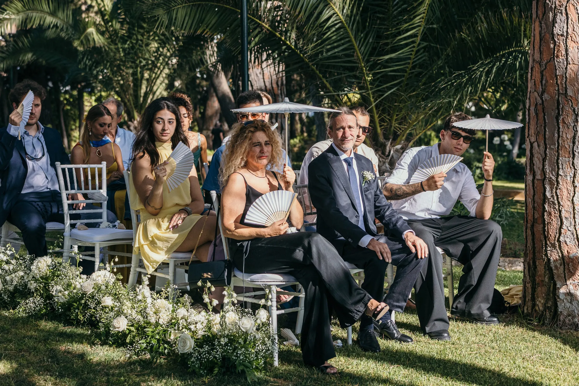 Guests in summer clothes seated in the shade of the trees for the garden ceremony