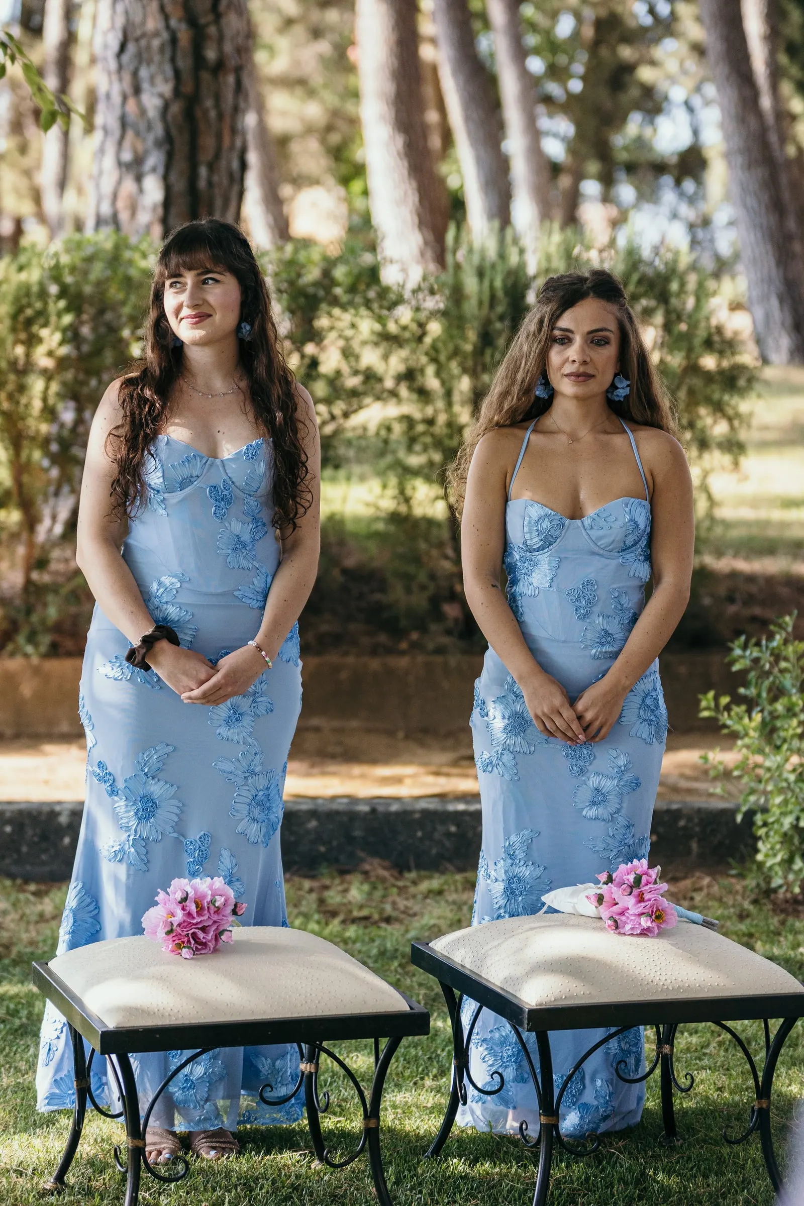 Two bridesmaids in matching blue floral dresses stand beside white stools in the garden