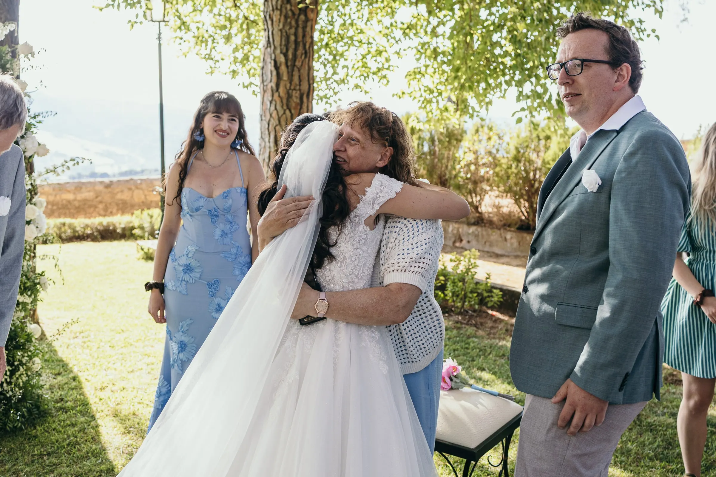 Sara is hugged by a guest on the lawn, a bridesmaid in blue beside them