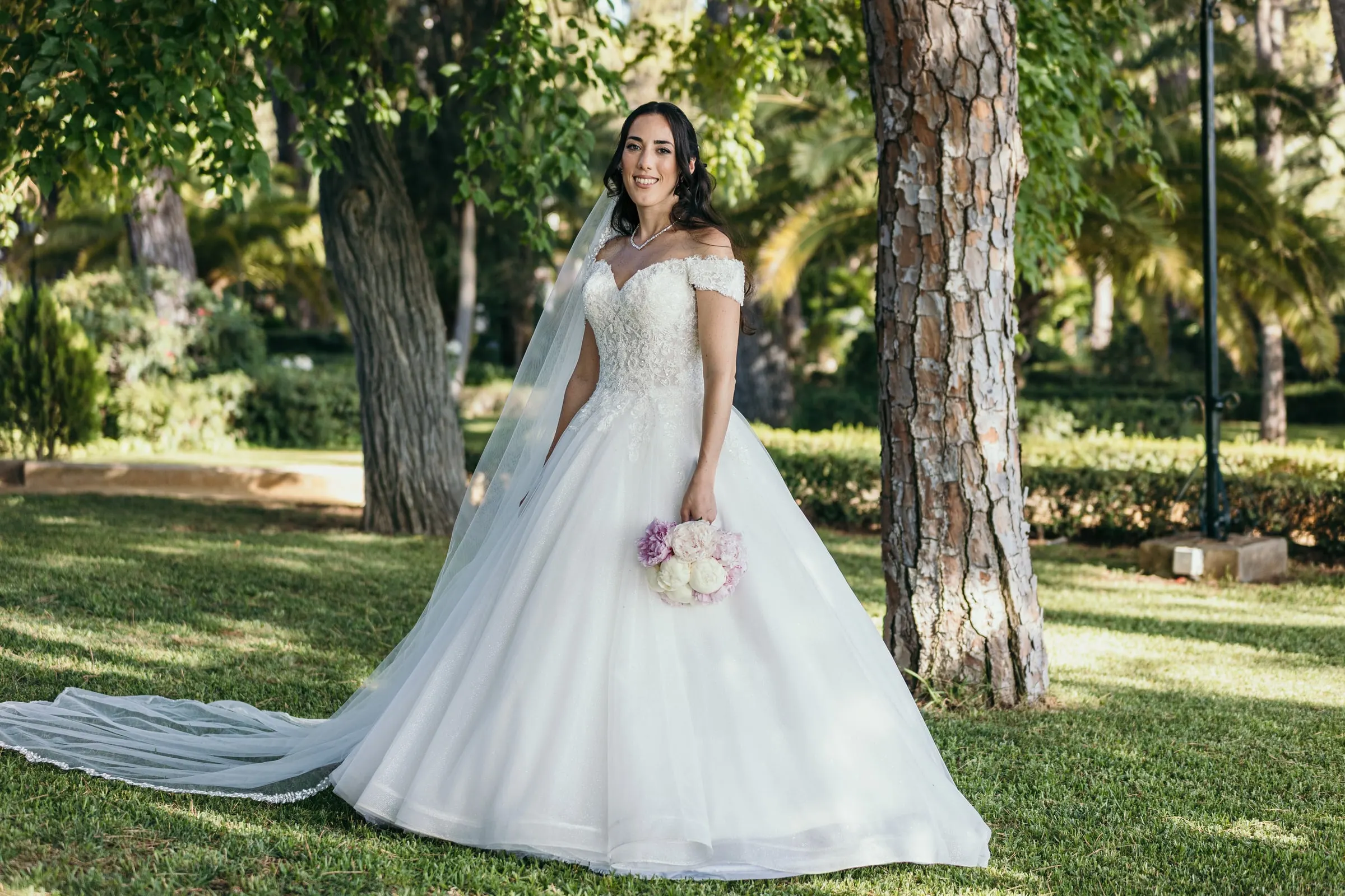 Sara holds her bouquet under the pines, her train spread across the grass
