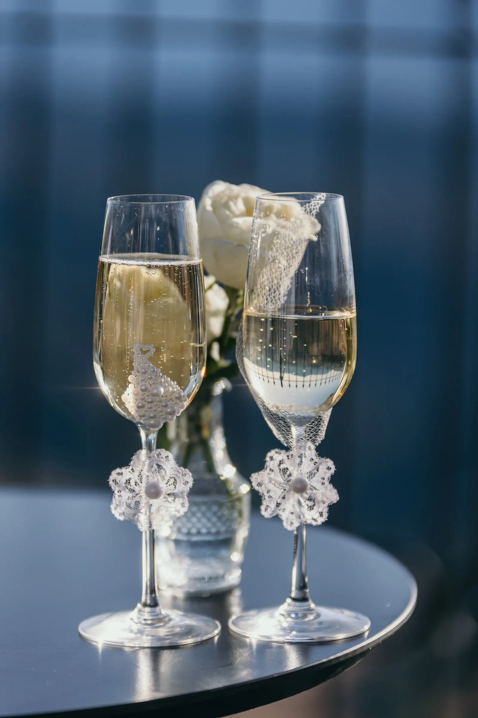 Two champagne flutes with lace details and a white rose on a dark table