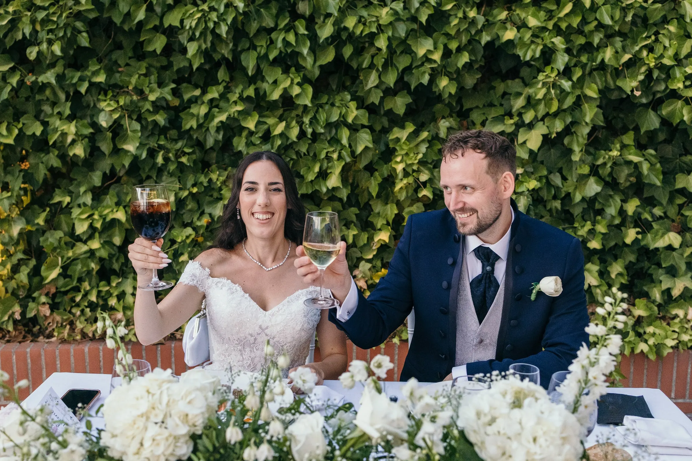 Sara and Vincent raise their glasses at the top table against a green hedge