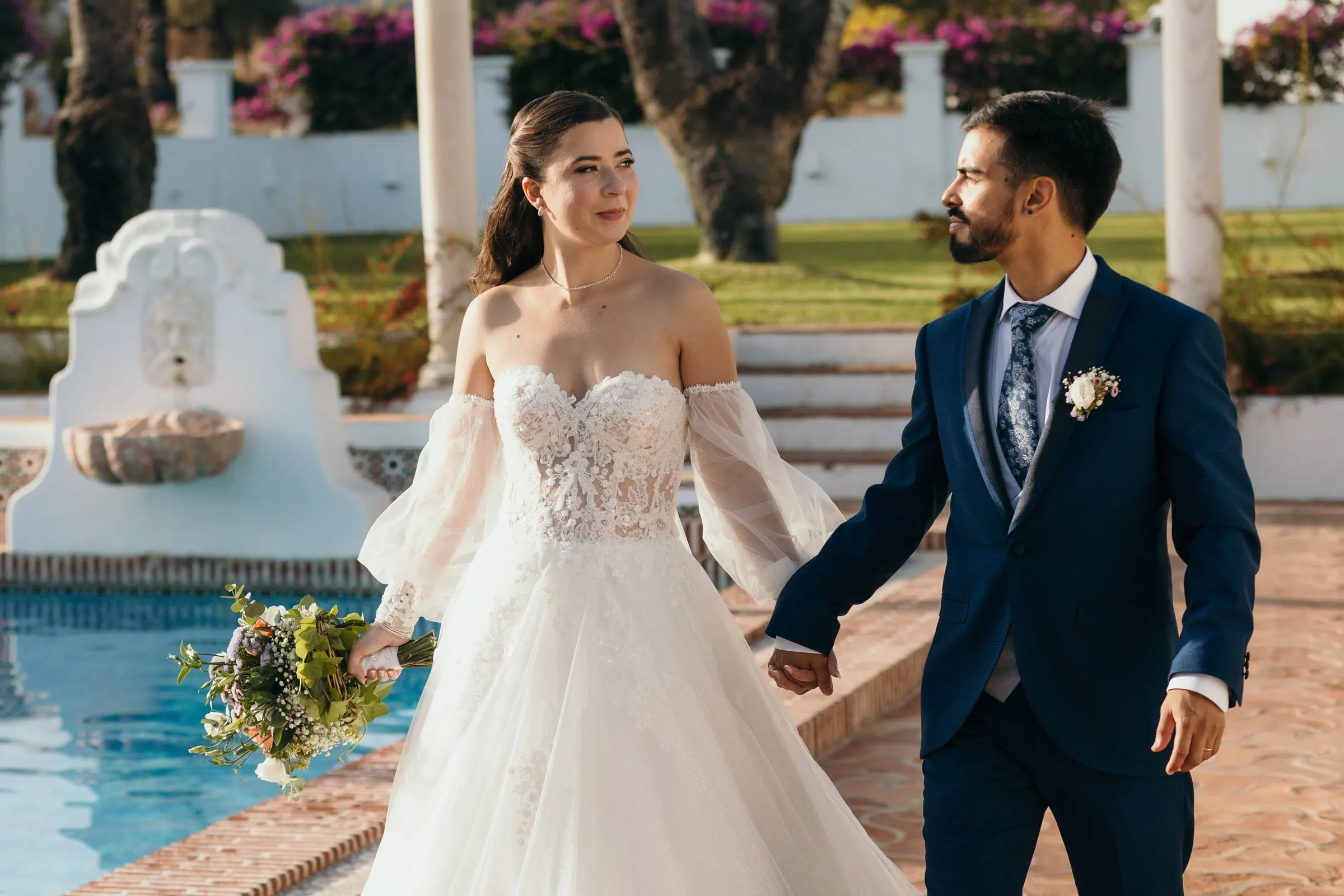 Bride and groom walking hand in hand past the pool and its tiled fountain, Cortijo San Francisco, Estepona