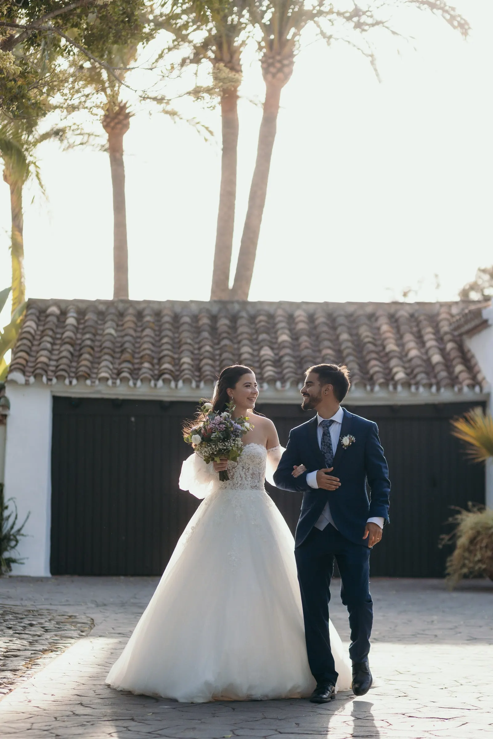 Bride and groom walking arm in arm across a courtyard under palm trees, Cortijo San Francisco, Estepona