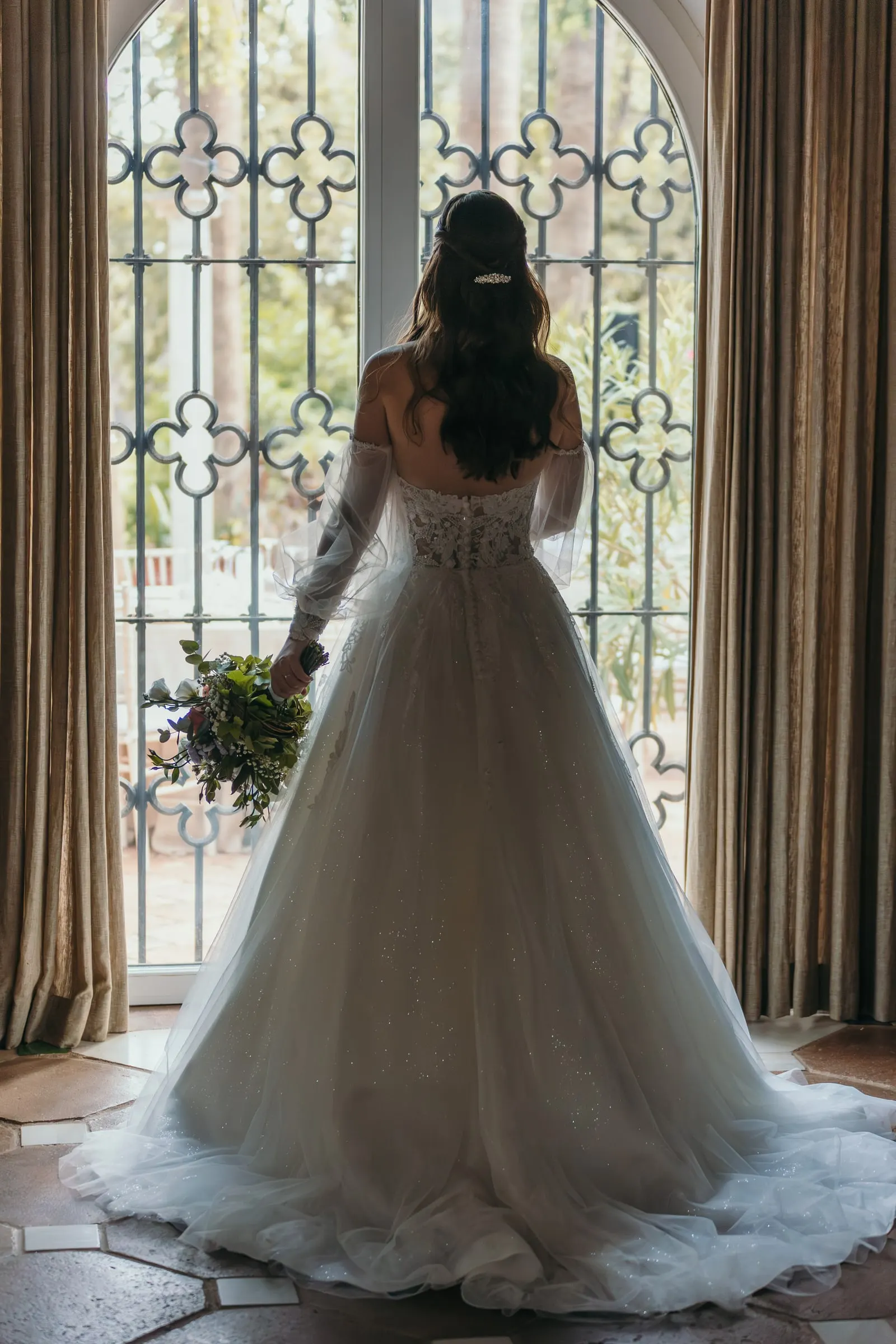 Bride seen from behind with her bouquet at a tall wrought-iron window, Cortijo San Francisco, Estepona