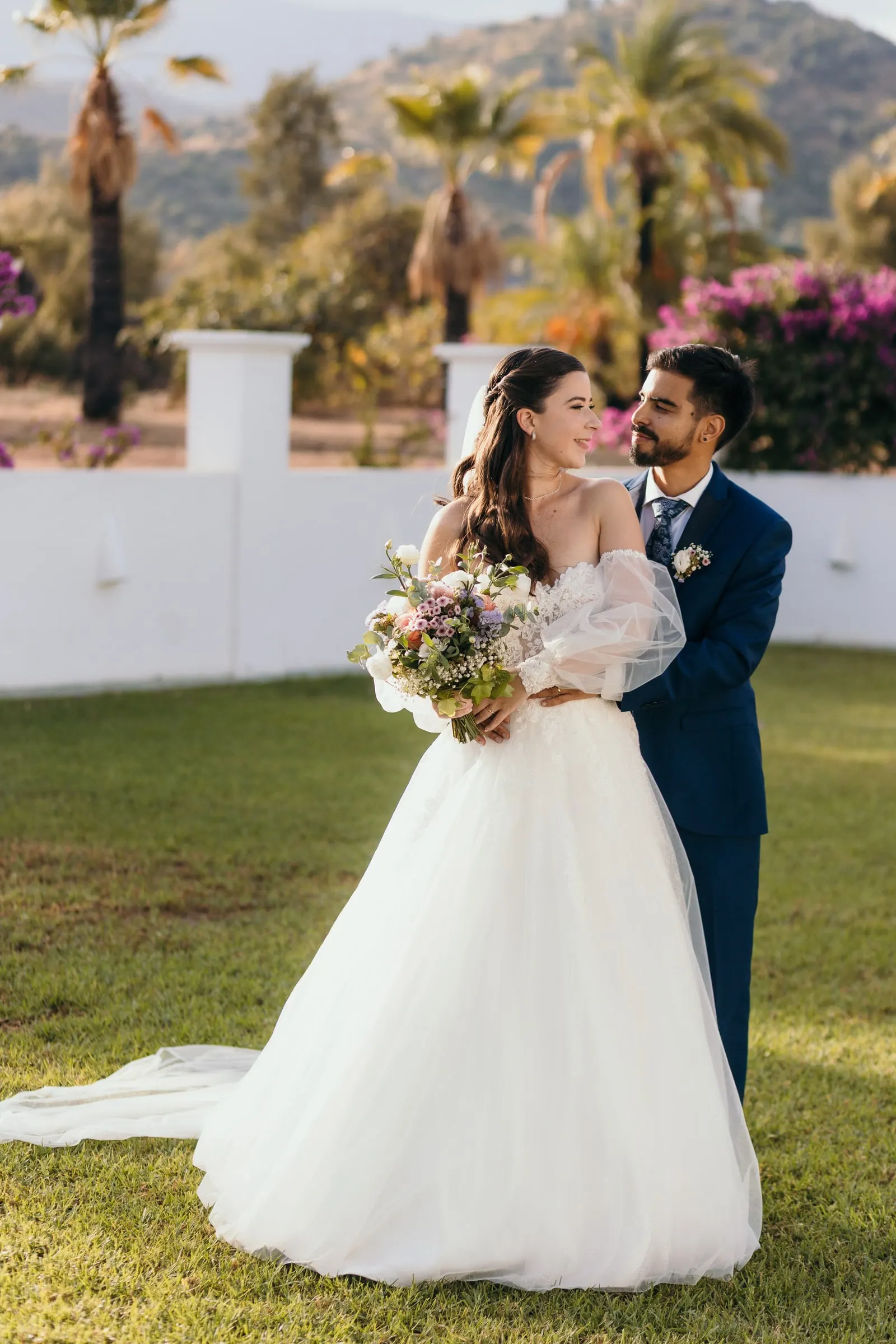 Groom hugging the bride from behind on the lawn with hills behind, Cortijo San Francisco, Estepona