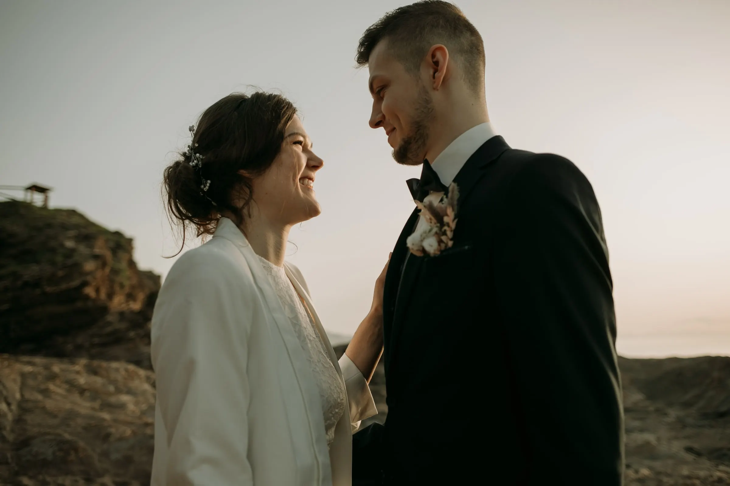 La novia ríe con el novio, con una caseta de madera en el promontorio detrás