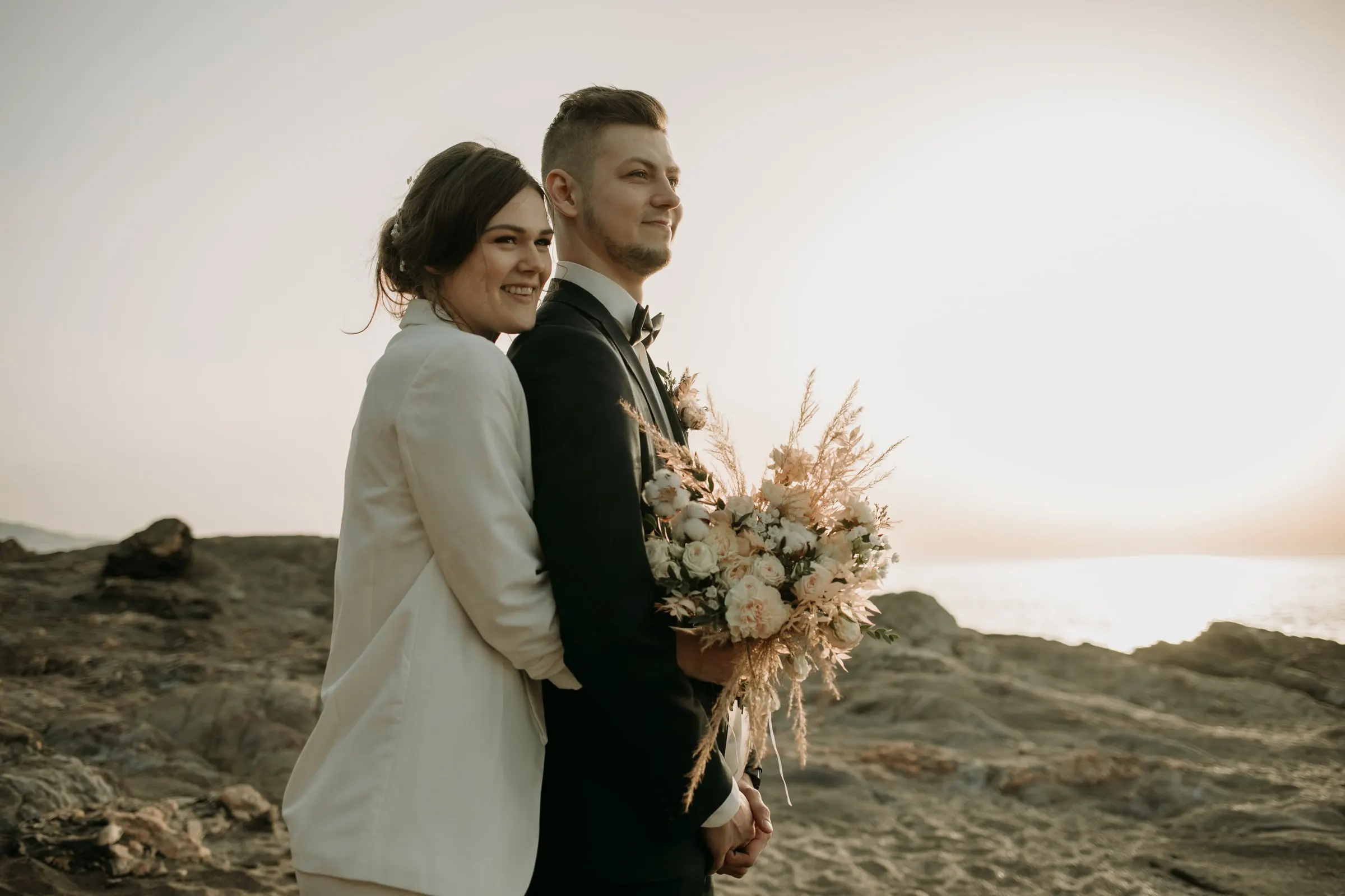 La novia abraza al novio por detrás con su ramo de flores secas y sonríe a cámara