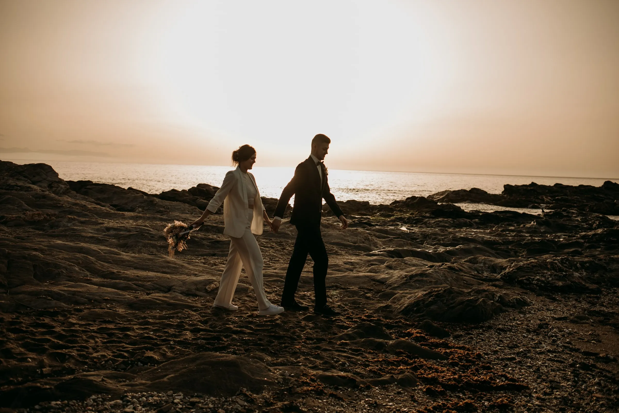 Los novios, pequeños ante el mar iluminado, cruzan la orilla rocosa
