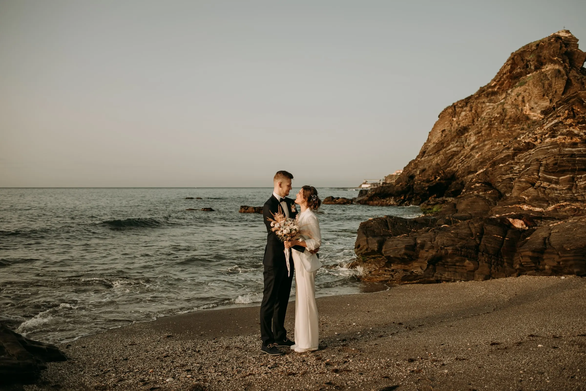 Bride and groom on a shingle beach beside a rocky headland, post-wedding session on the Costa del Sol