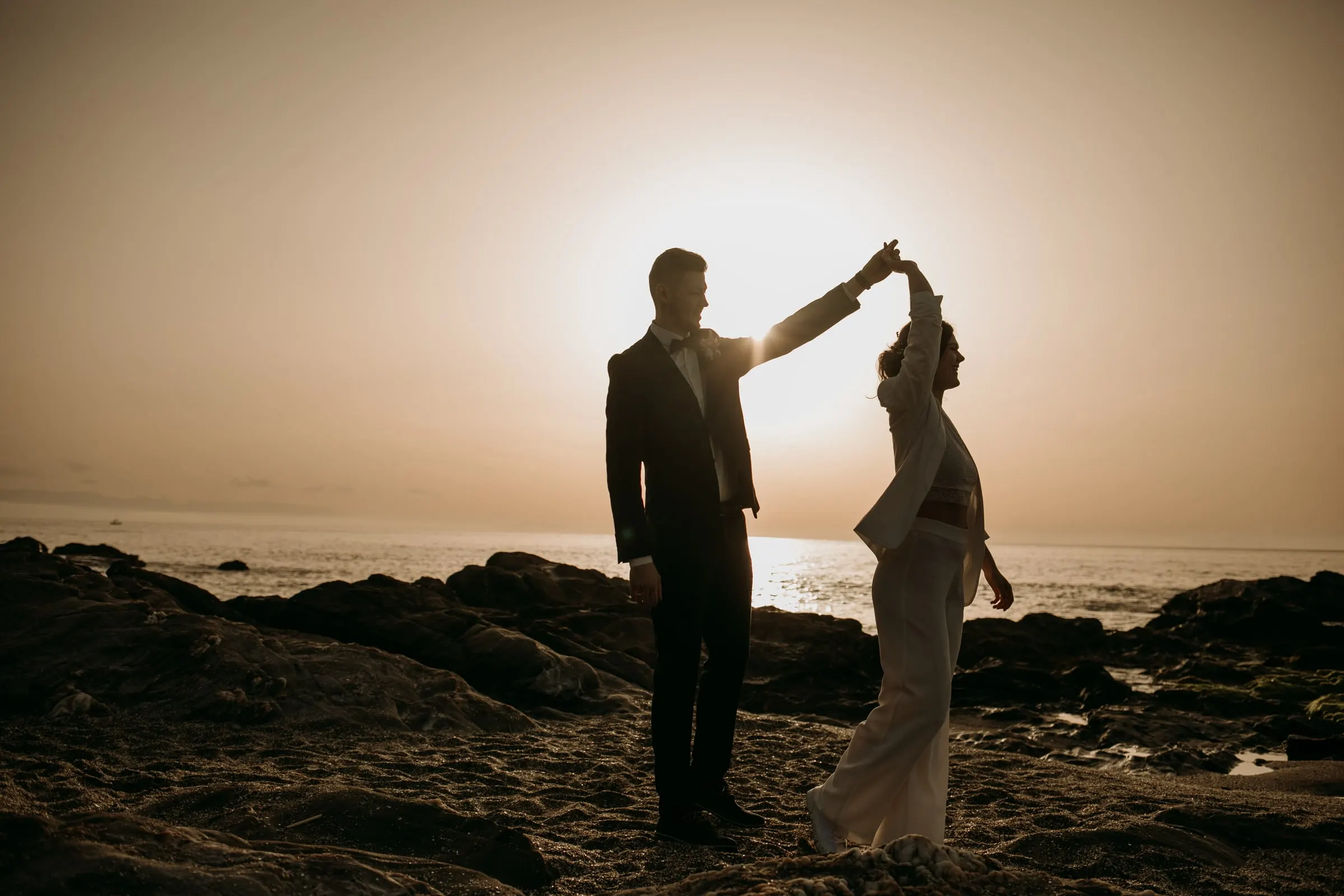El novio hace girar a la novia a contraluz sobre las rocas al atardecer