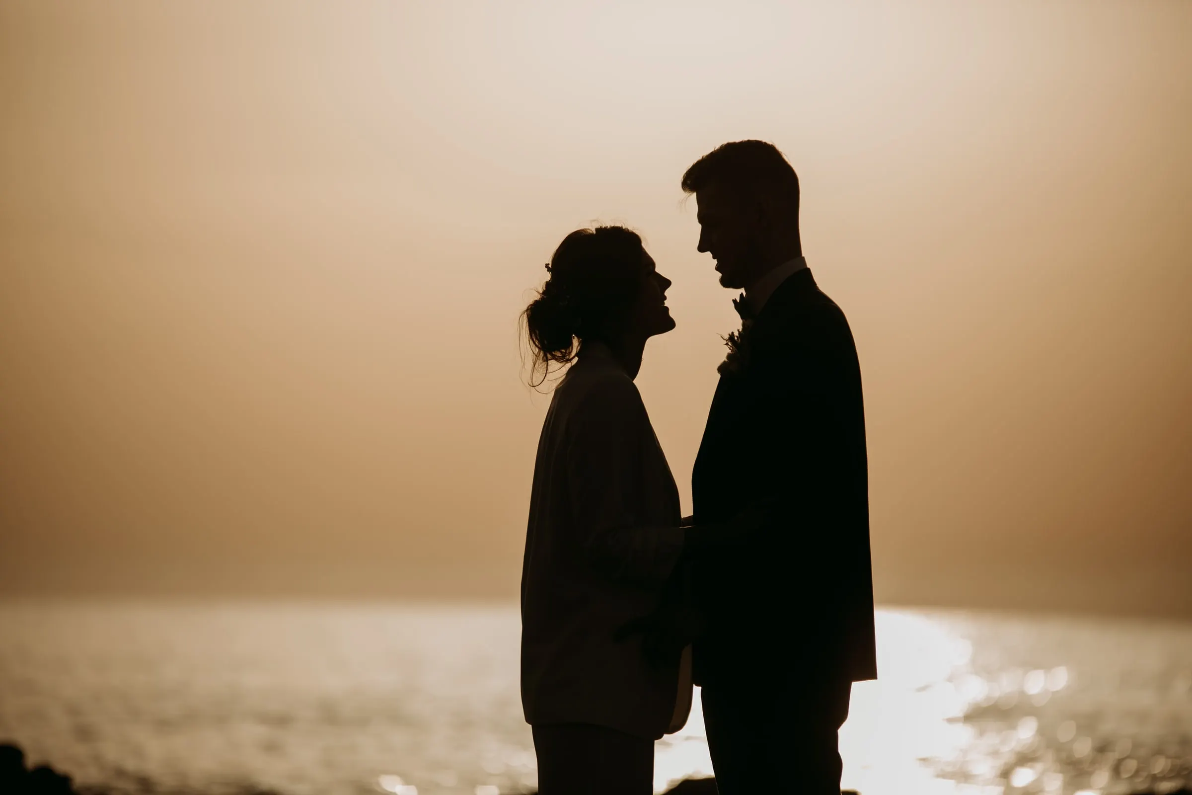 Siluetas de los novios frente a frente, con el sol destellando en el agua