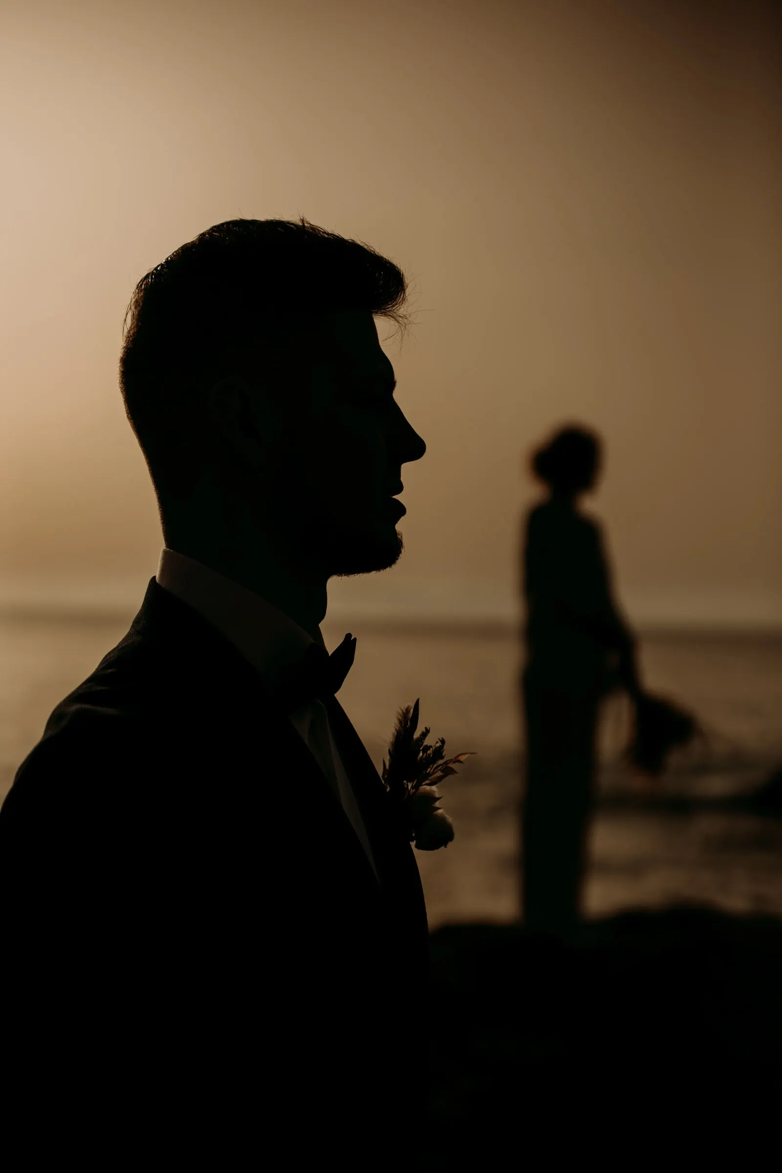 El novio a contraluz con la novia desenfocada junto al mar