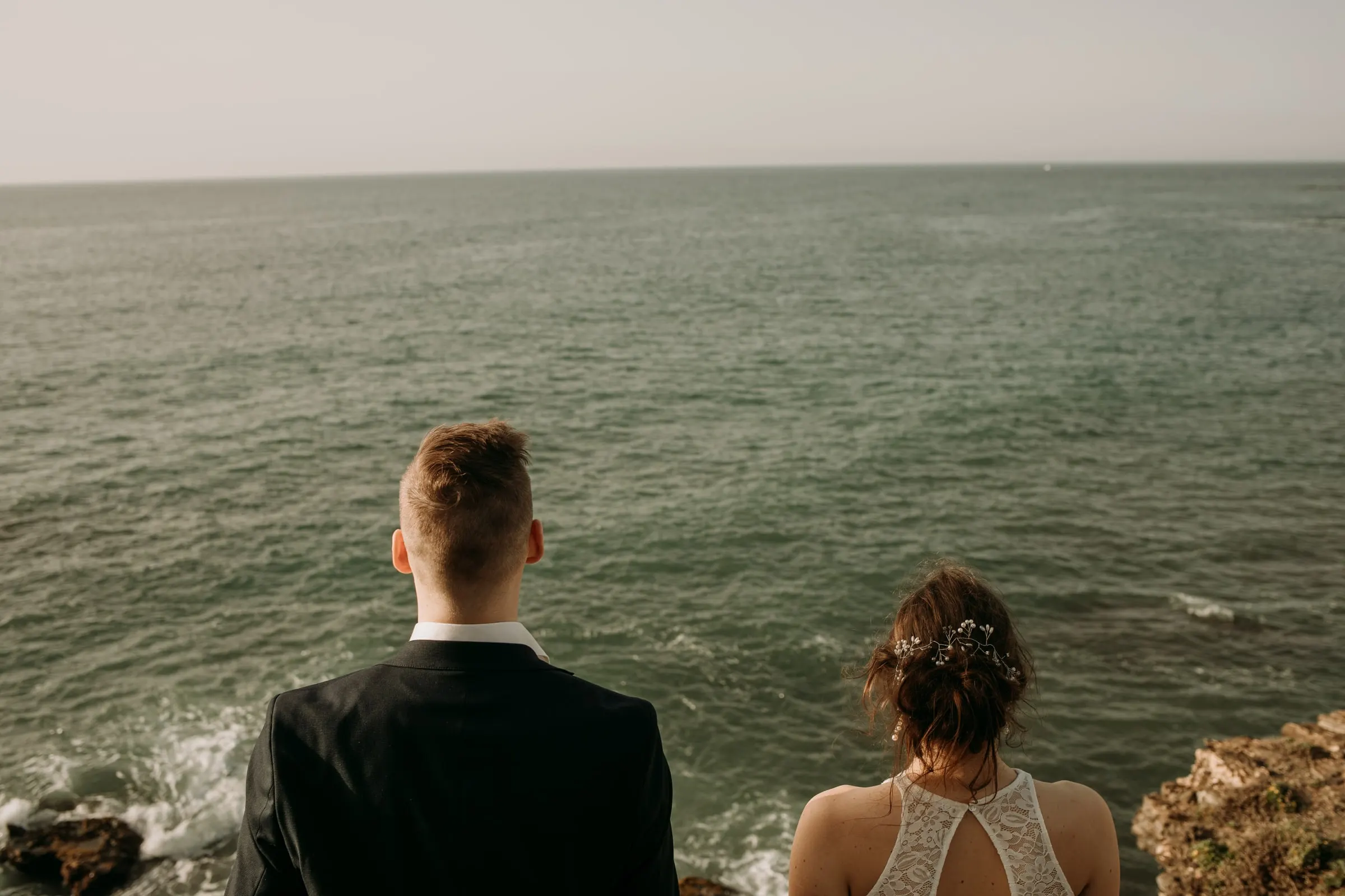 Los novios de espaldas miran el mar abierto desde el acantilado, ella con un top de encaje con la espalda al aire