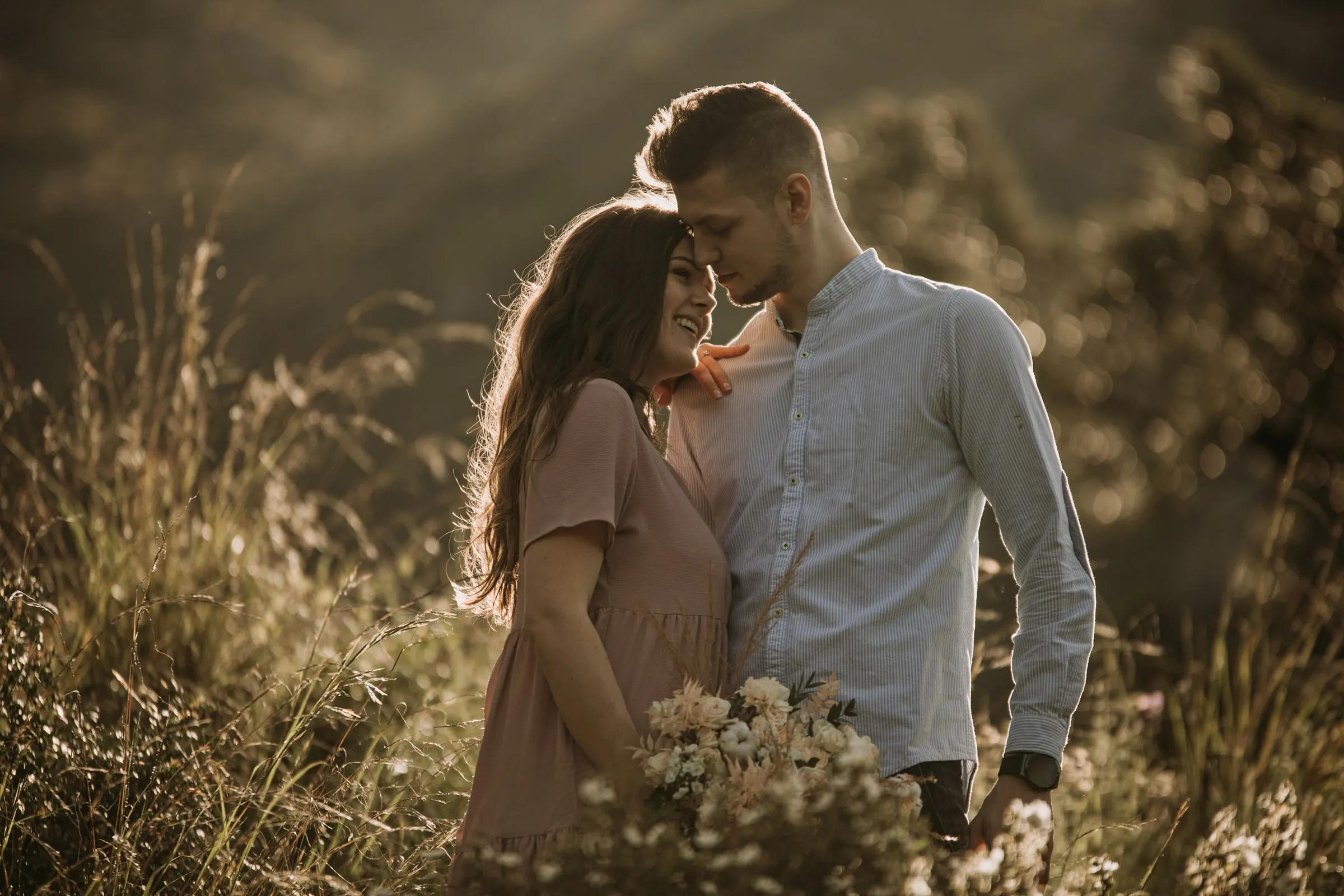 La futura novia con vestido rosa se apoya en su prometido con los ojos cerrados entre la hierba dorada