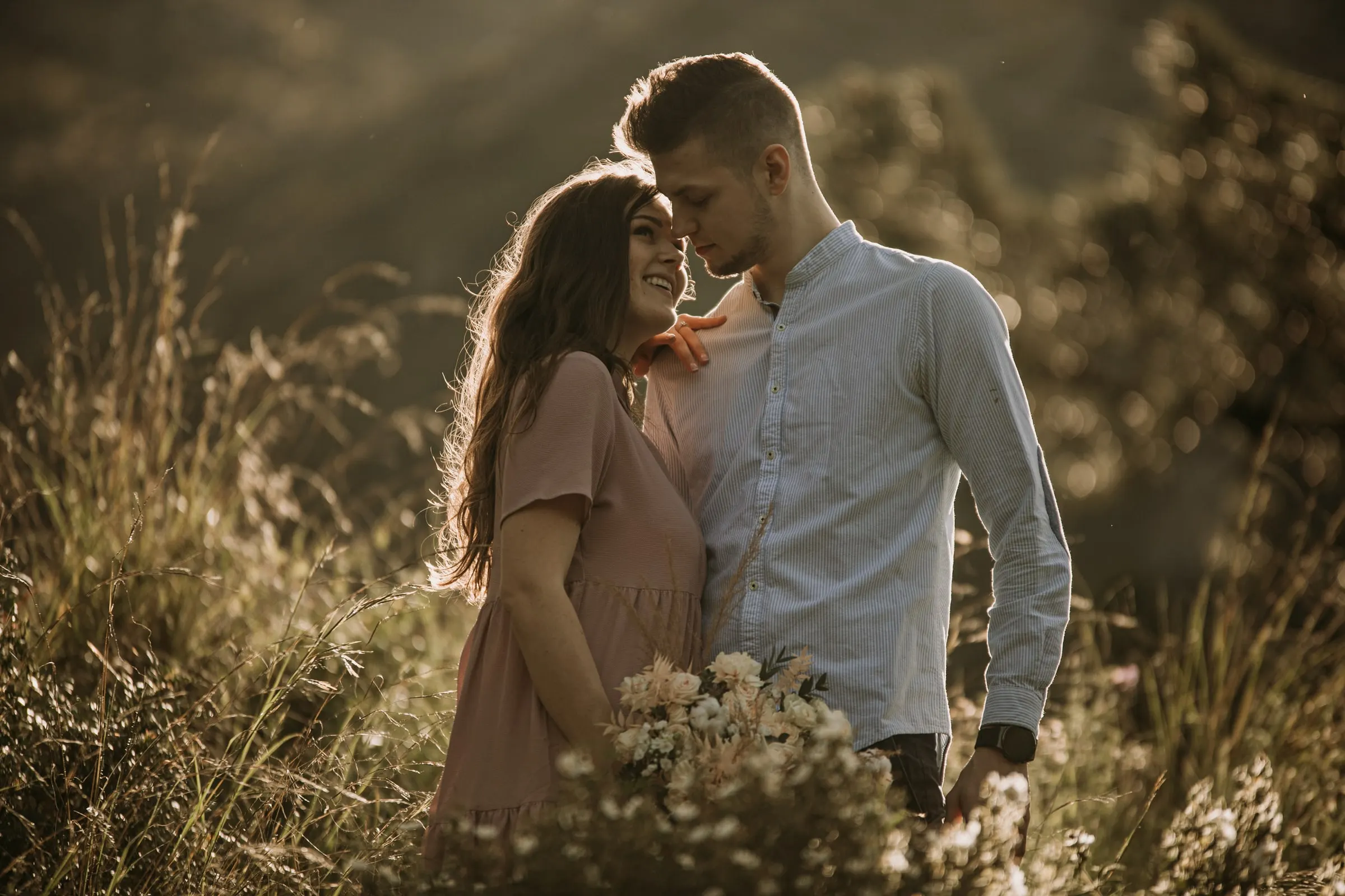 La pareja se sonríe frente a frente, con el pelo de ella iluminado por el sol y un ramo silvestre delante