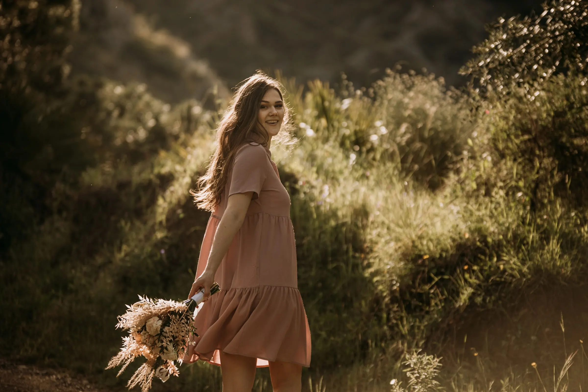 La futura novia con vestido rosa camina entre la hierba alta y mira atrás