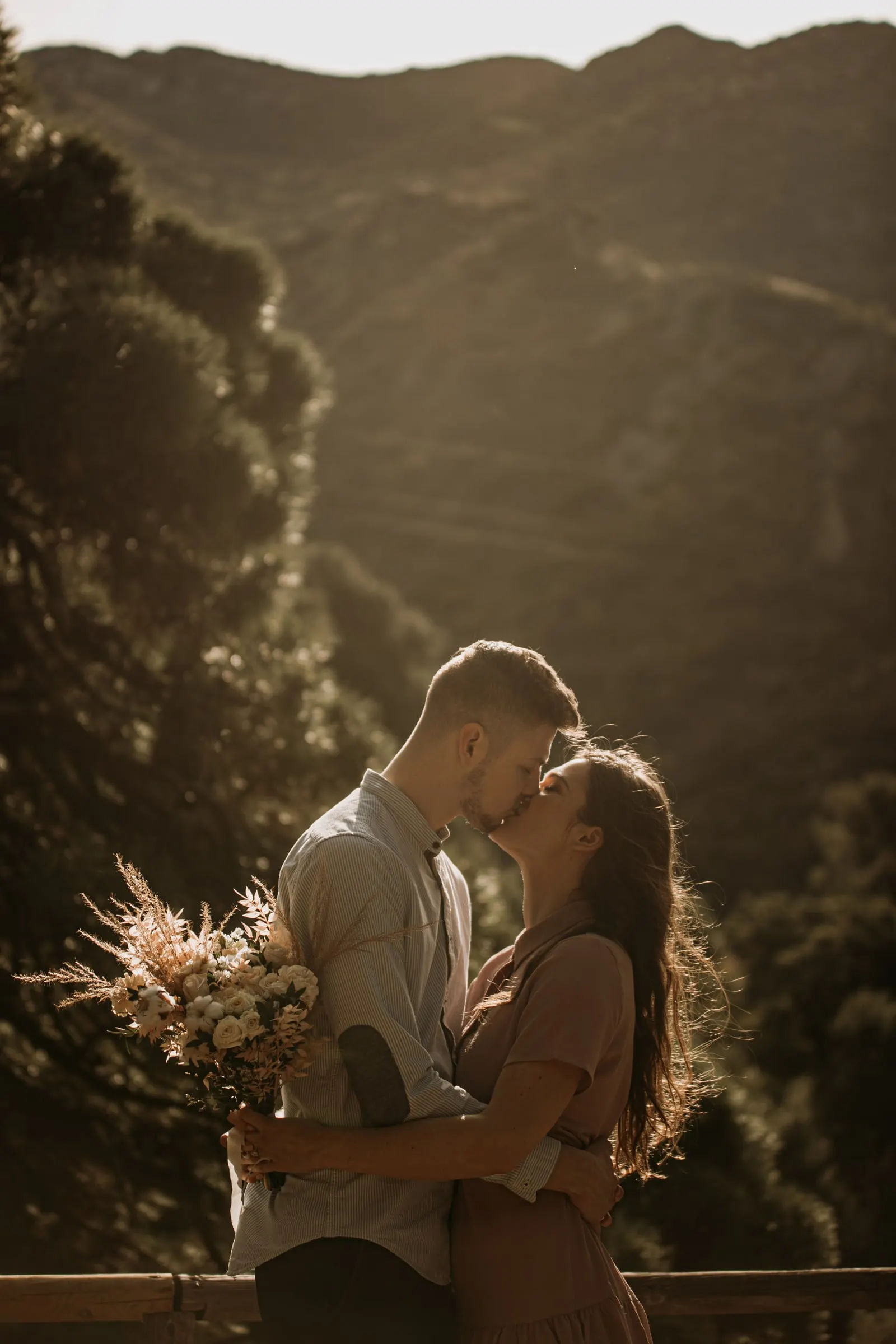 Beso de la pareja junto a una barandilla de madera, con pinos y montes en luz suave