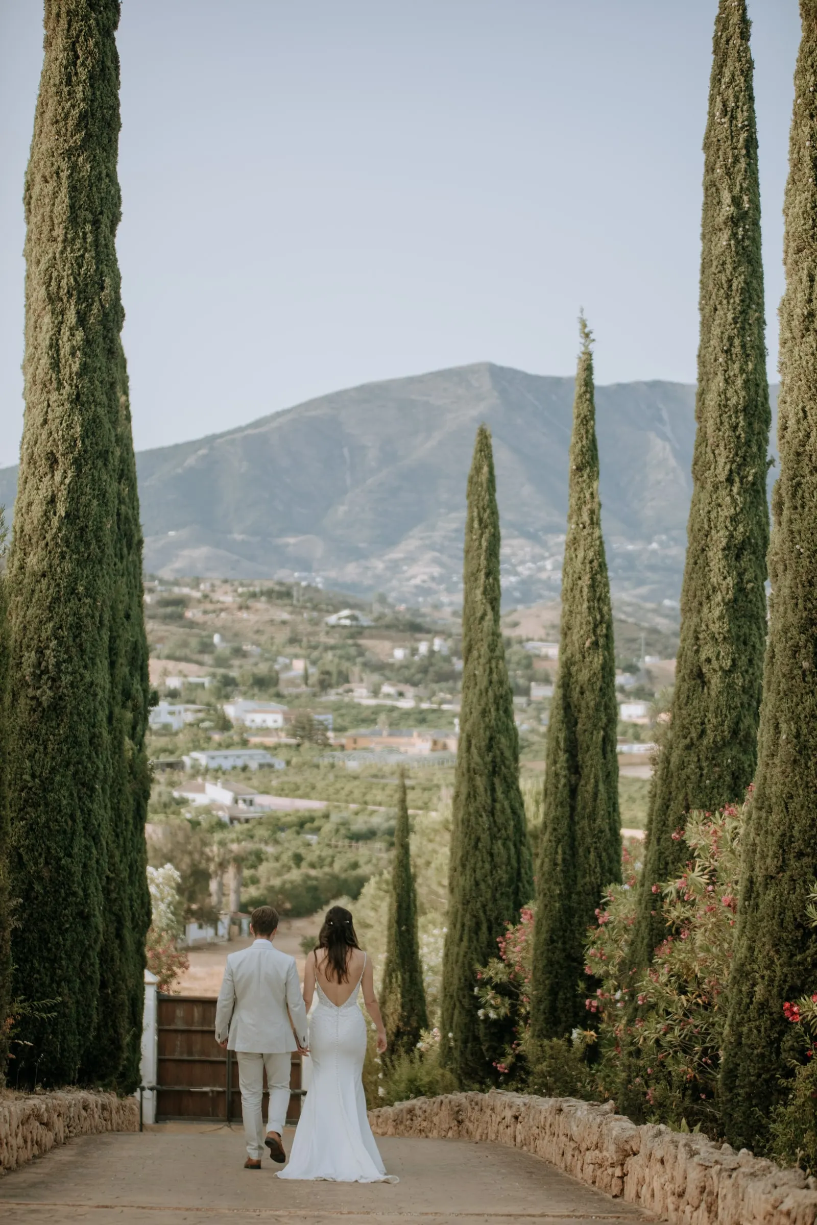 Bride and groom walking down a cypress-lined path with the mountains behind, wedding in Málaga