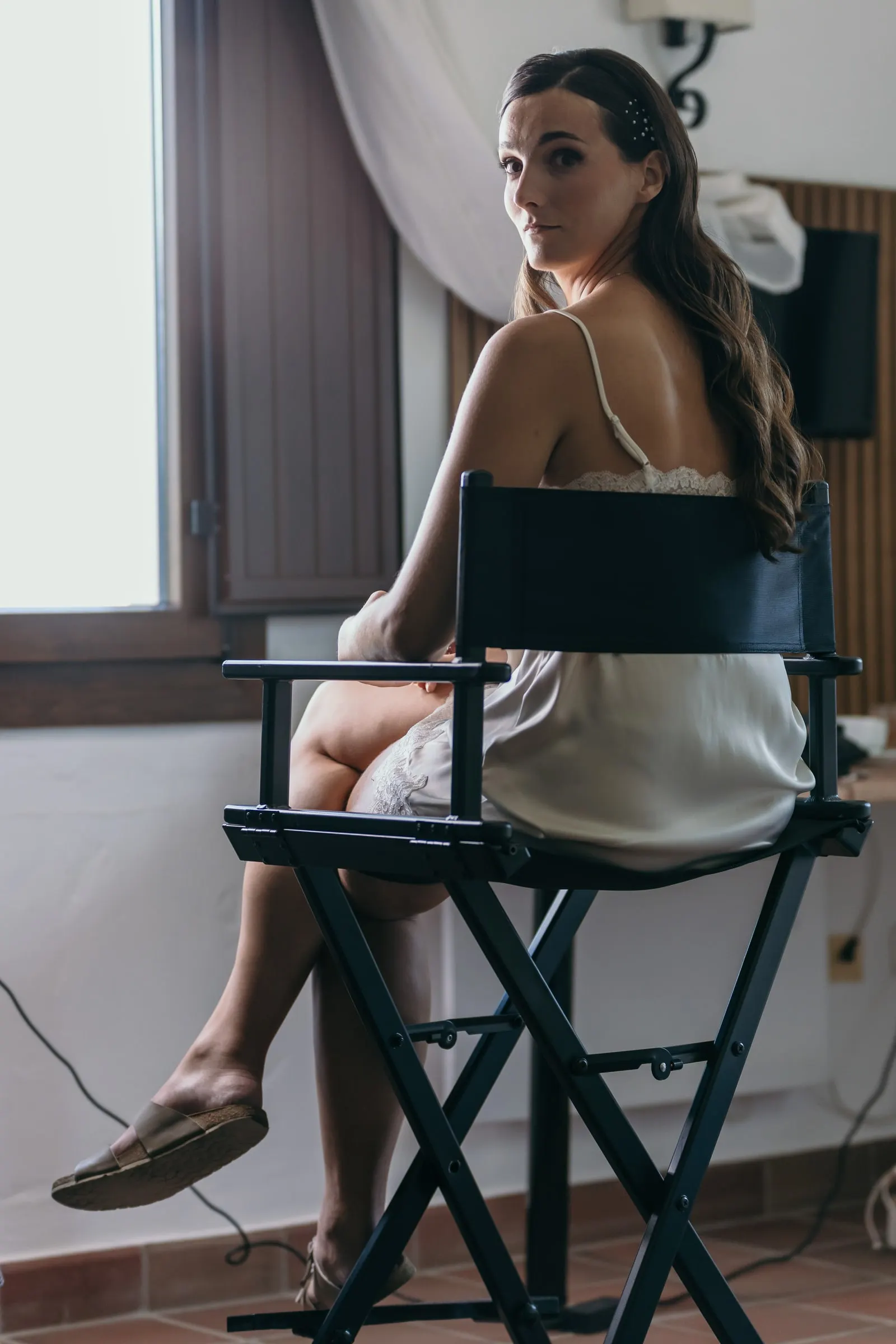 Bride in a silk slip looking over her shoulder from a director's chair by the window