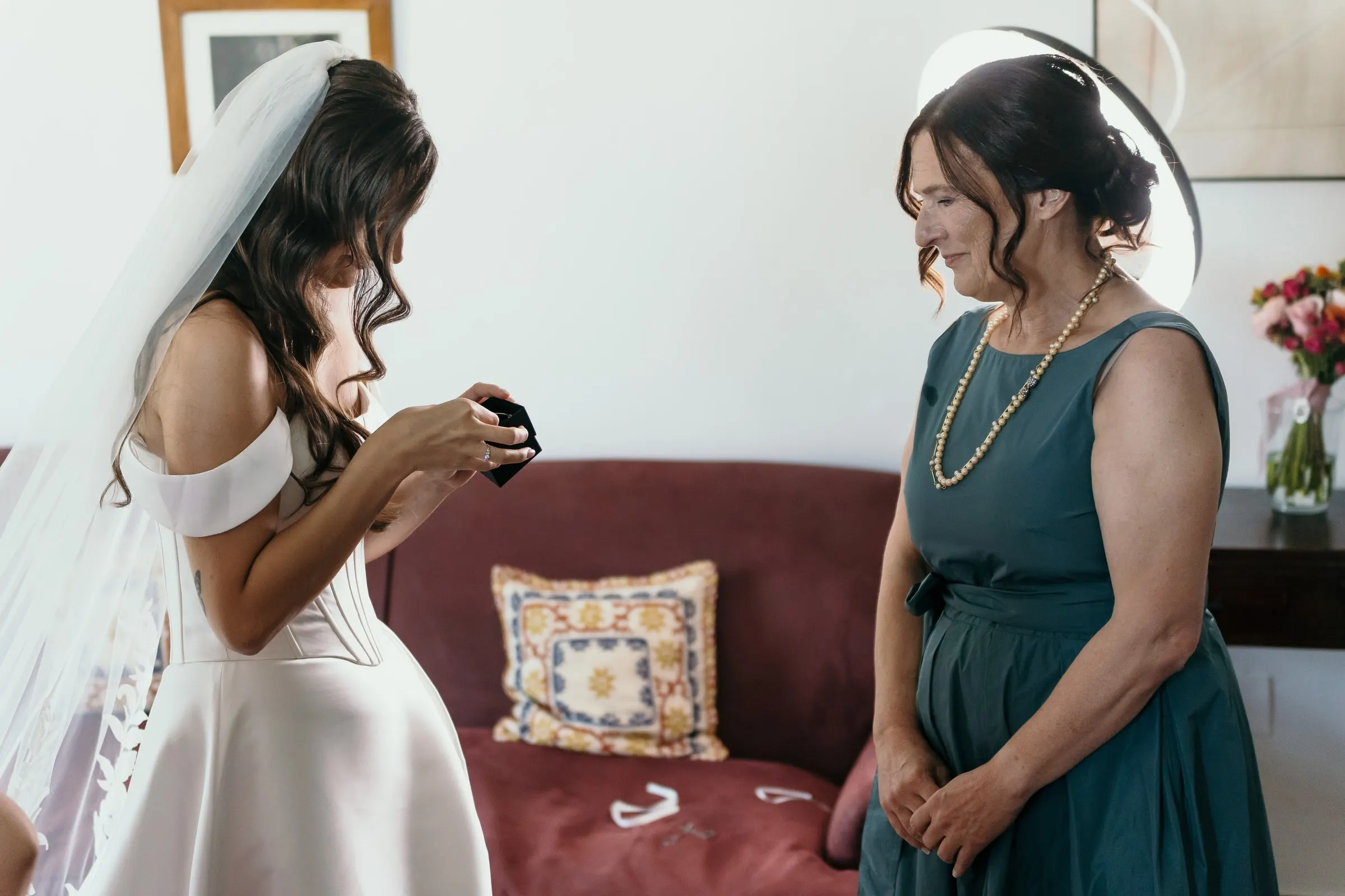 Bride opening a small gift box in front of a red sofa while the woman in teal watches