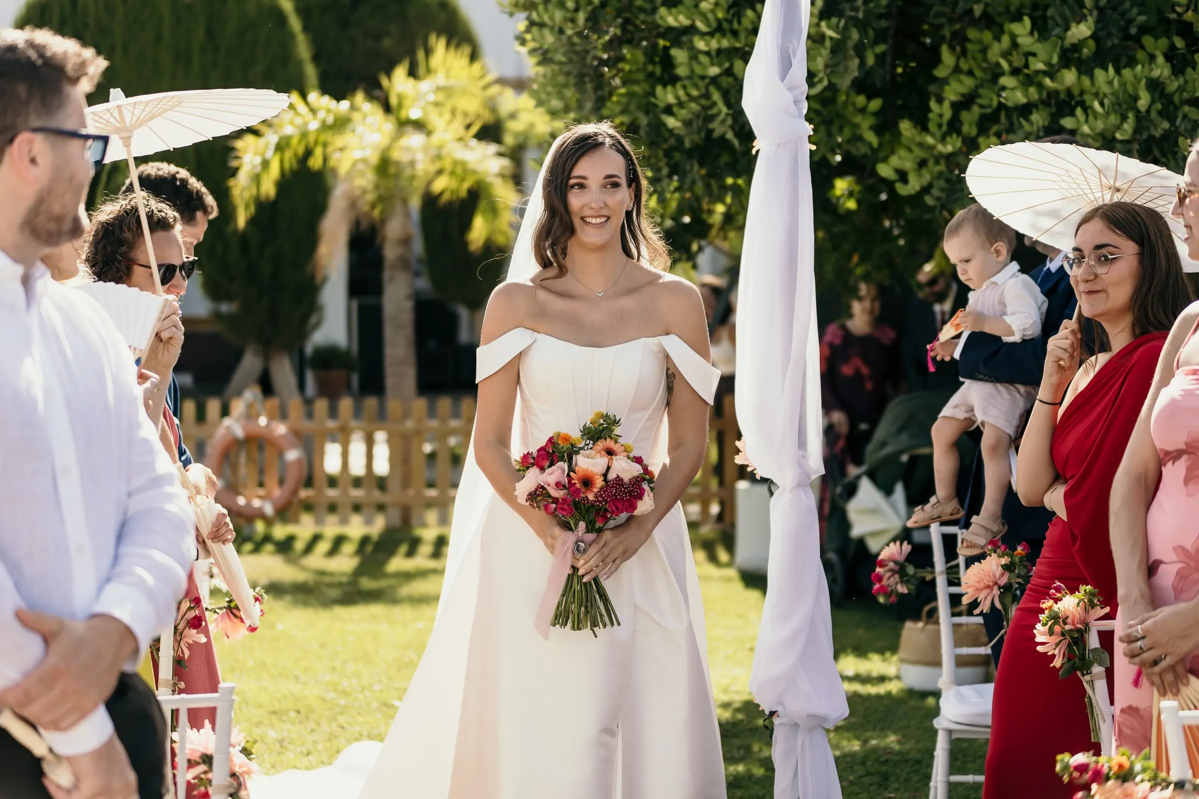 Bride smiling down the grass aisle between guests with paper parasols and a baby