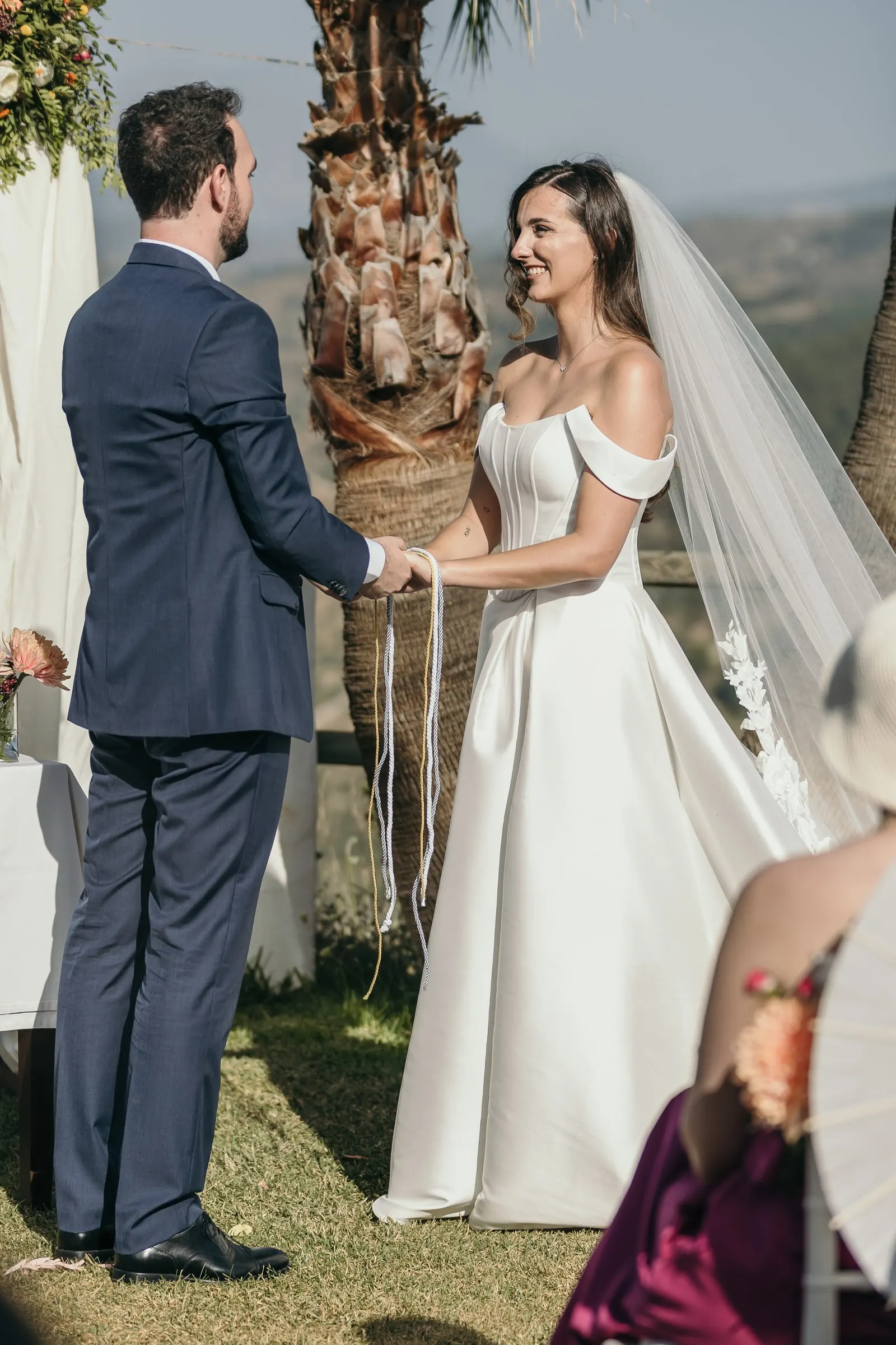 Bride and groom facing each other with joined hands beside a palm tree, hills behind
