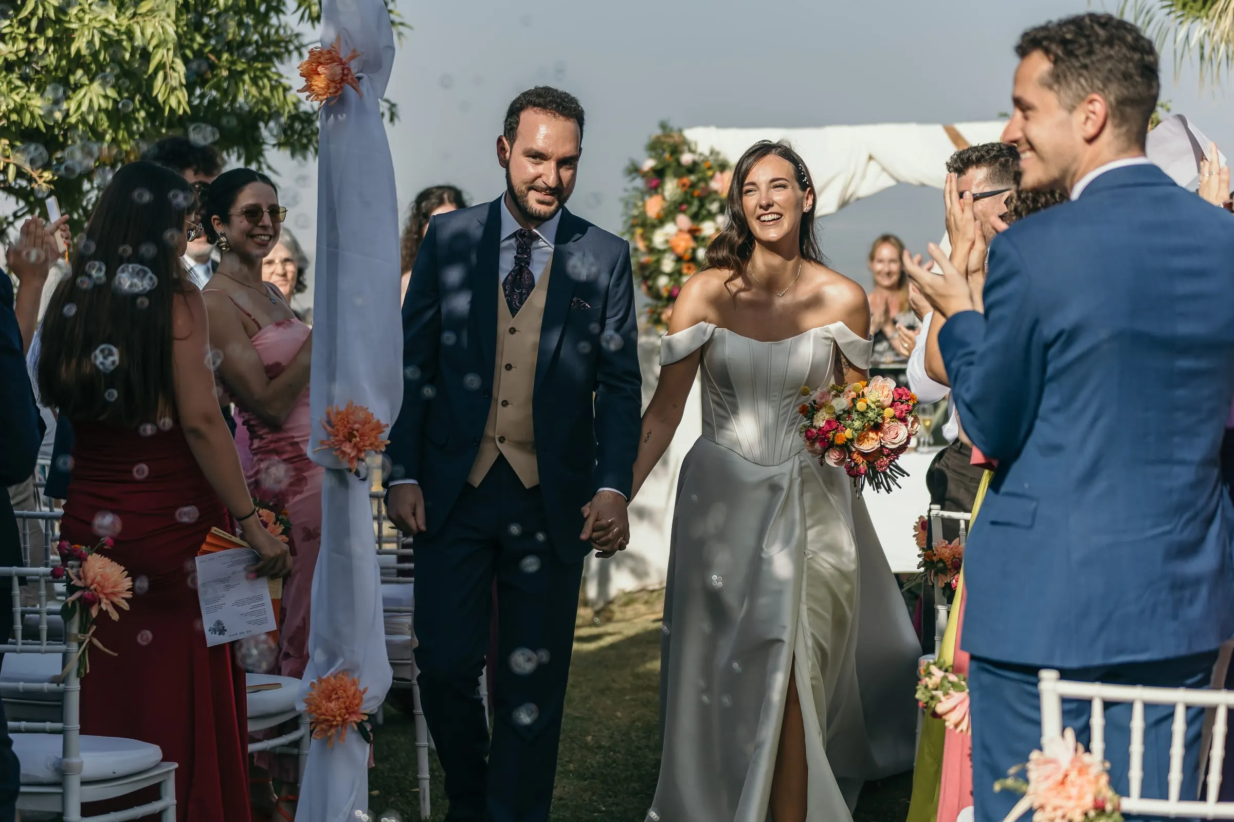 Bride and groom walking back up the aisle among soap bubbles and cheering guests