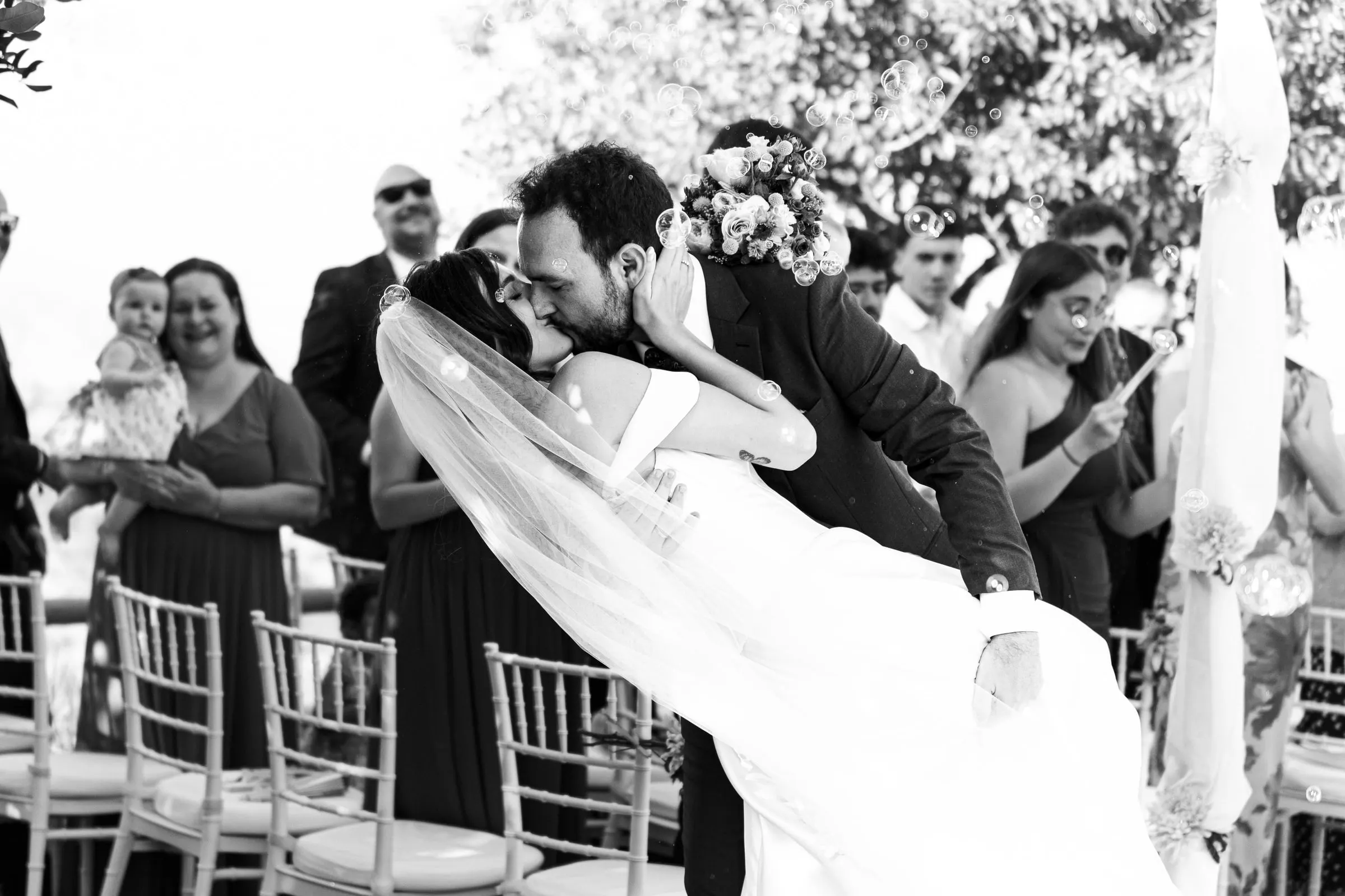 Black and white of the groom dipping the bride for a kiss as guests cheer