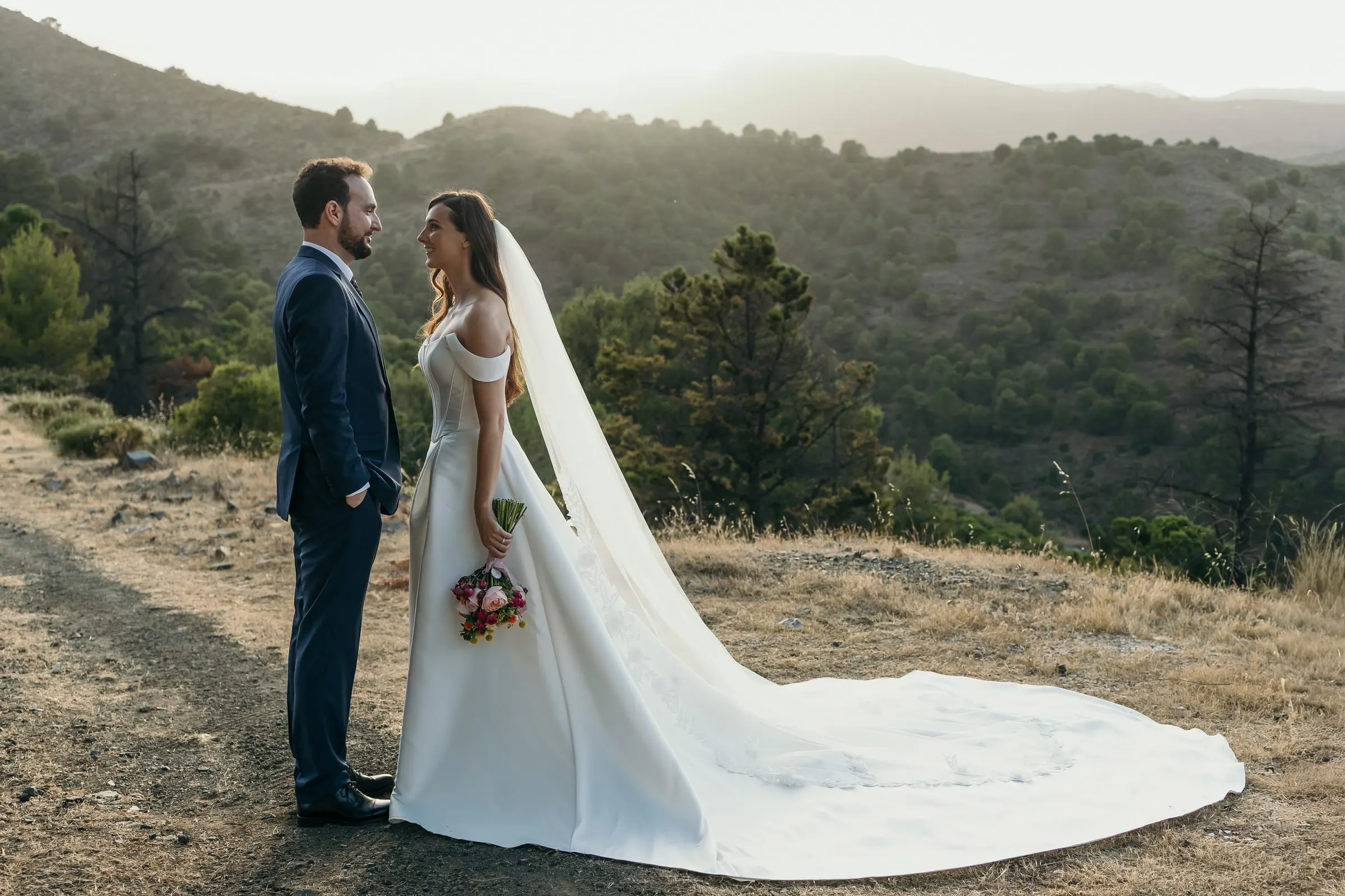 Bride and groom facing each other on a hilltop track with layers of mountains behind, wedding on the Costa del Sol